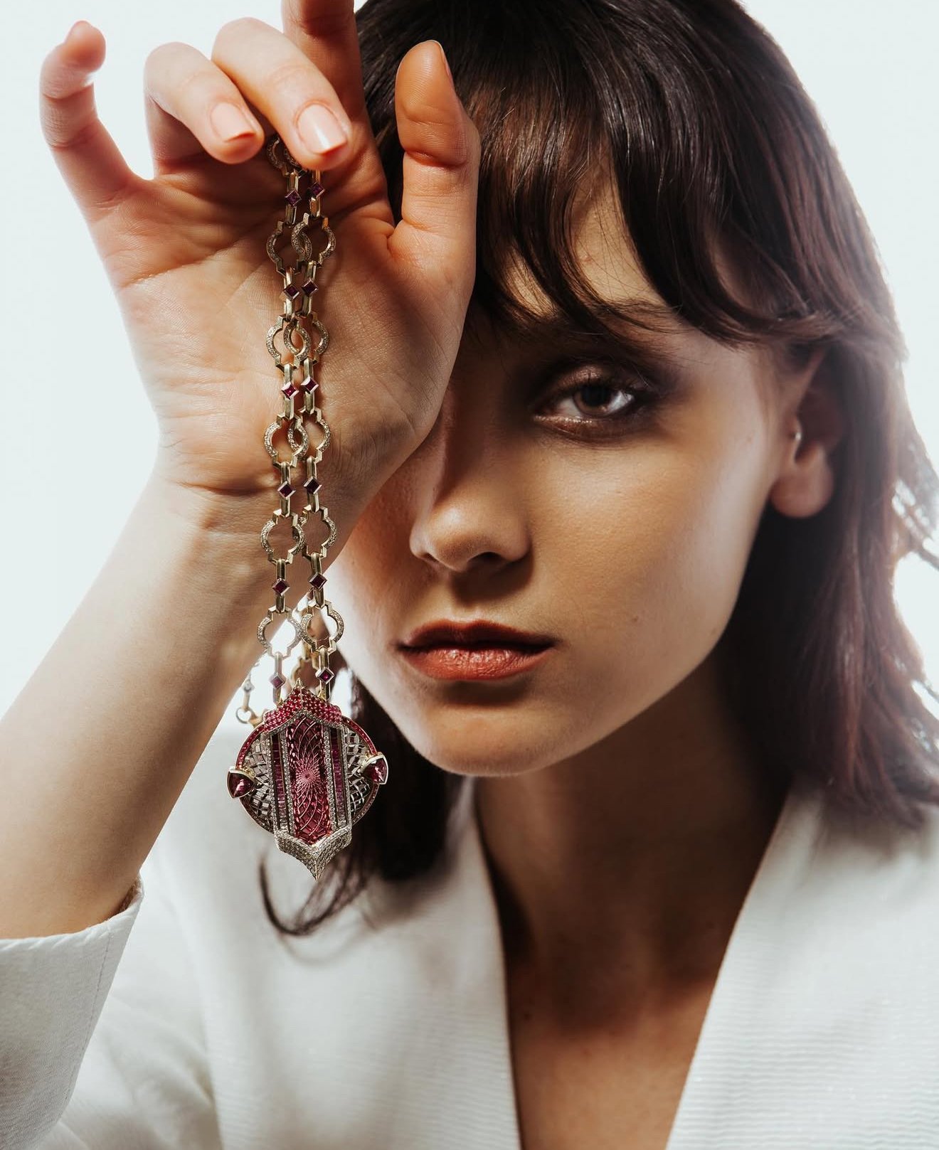 A woman with reddish-brown hair and light skin holding a large, ornate pink and silver pendant necklace near her face, with a neutral background.