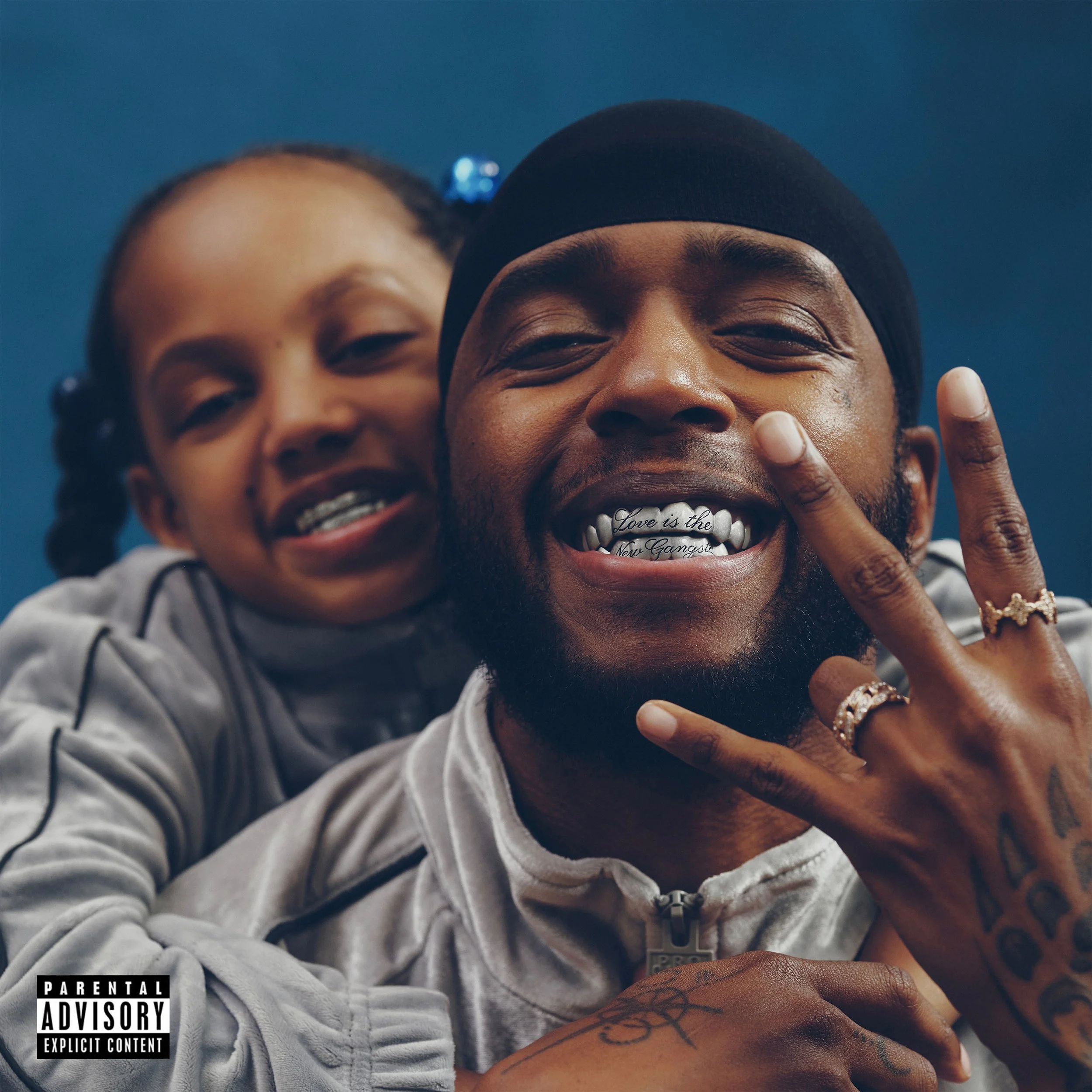 Close-up of two smiling people, a girl on the left and a man on the right, with the man making a hand sign. The man has a tattooed arm and is wearing a black durag and grillz that say "Love is the New Gangsta." 