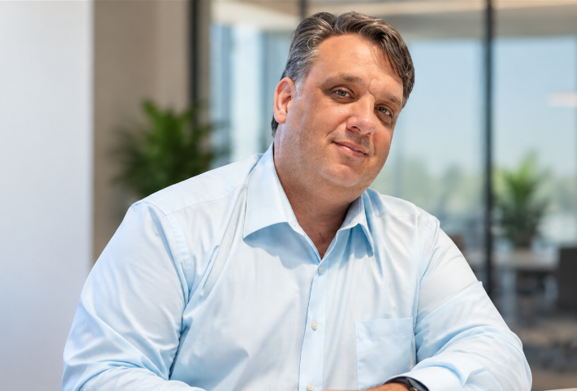 A middle-aged man with dark hair, wearing a light blue dress shirt, sitting in a modern office with large windows and potted plants in the background.