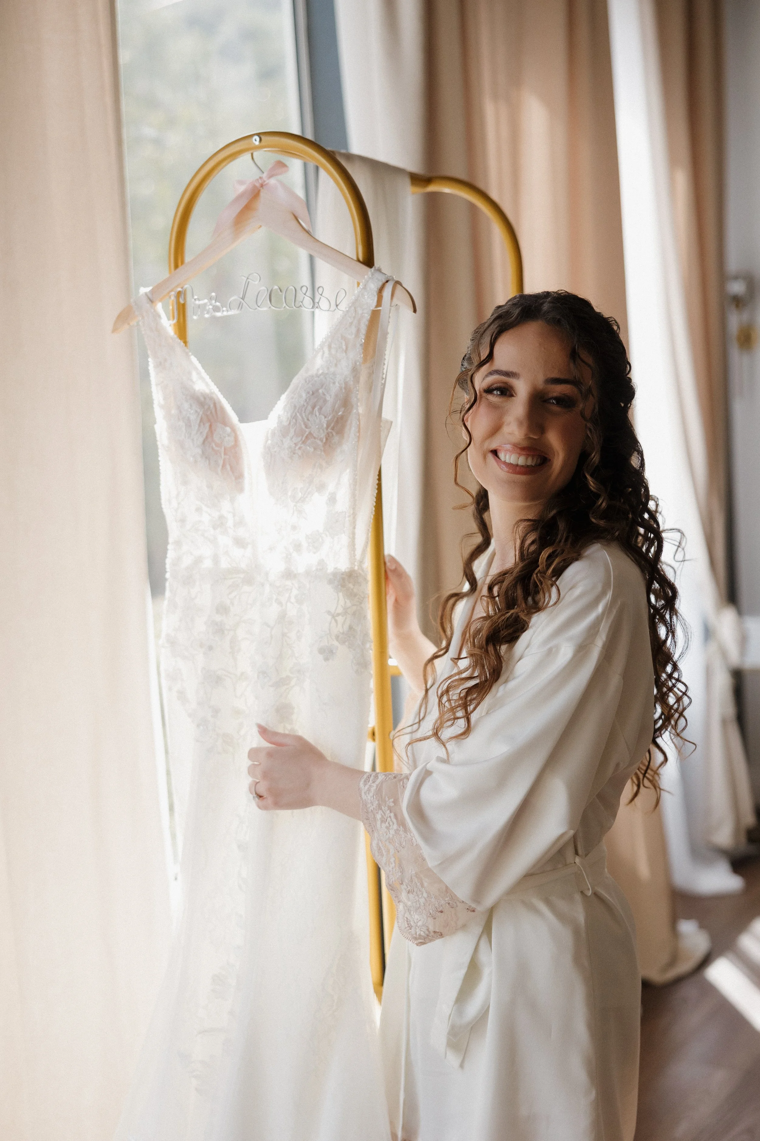 Bride getting ready for wedding standing next to her wedding dress on a rack and smiling
