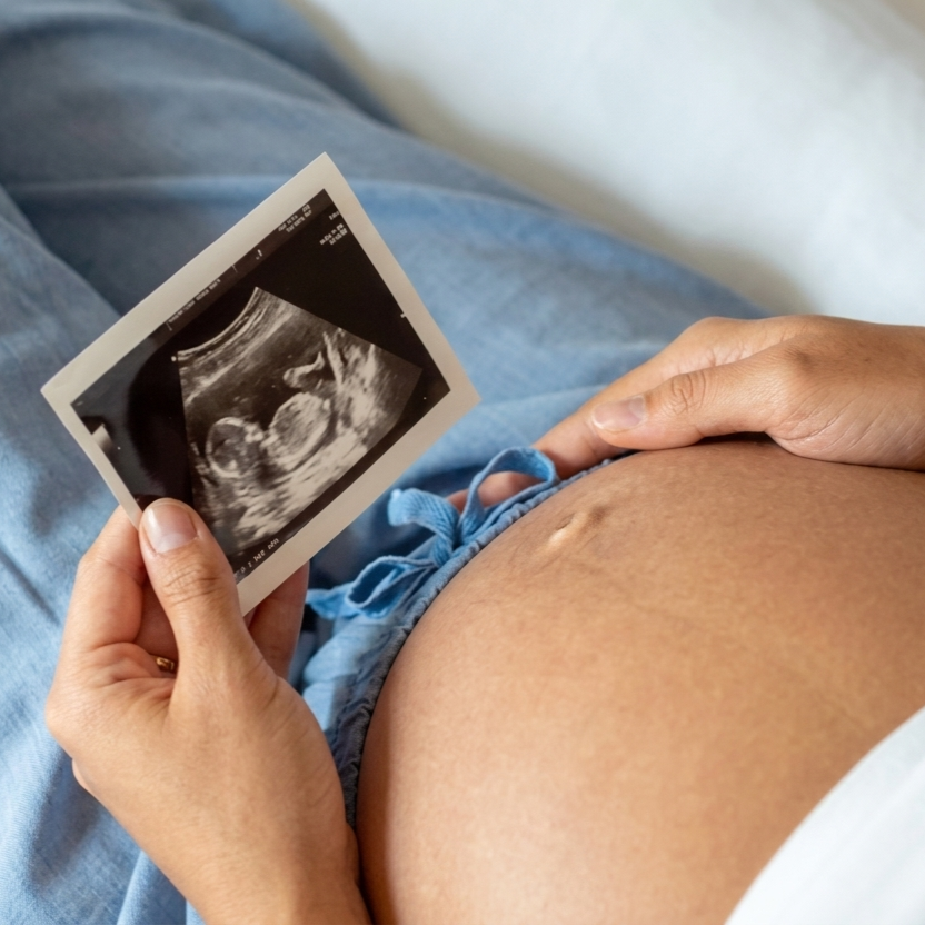 Person holding an ultrasound image while resting with a hand on their pregnant belly.