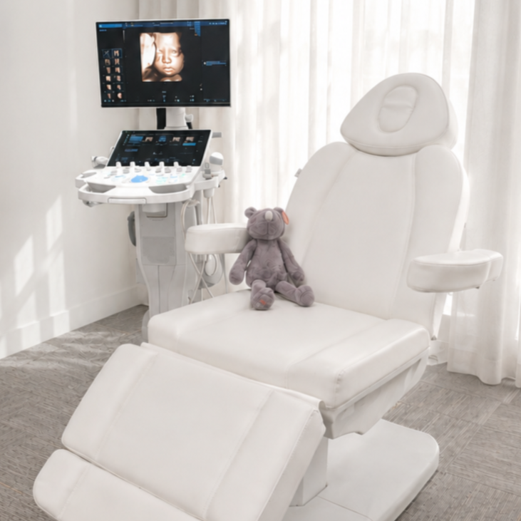 Medical examination room with an ultrasound machine, a white examination chair with a gray teddy bear sitting on it, and a monitor displaying a 3D ultrasound image of a fetus.
