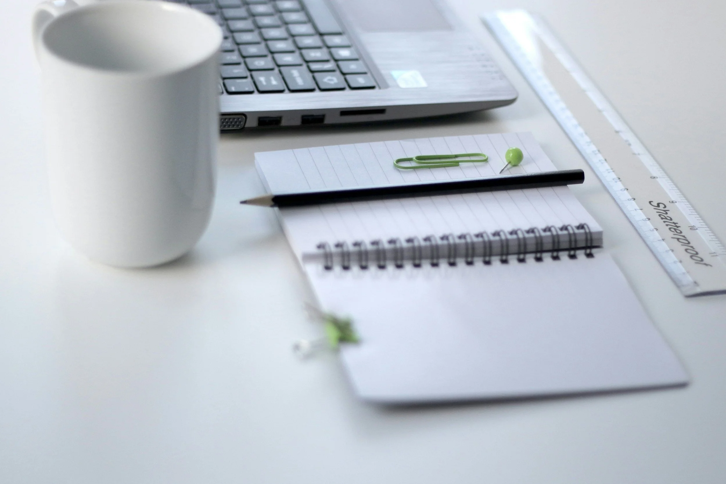 Close-up of a desk with a gray laptop, a white coffee mug, a green paperclip, a green push pin, a black pen, a spiral notebook, a lined notepad, and a transparent ruler.