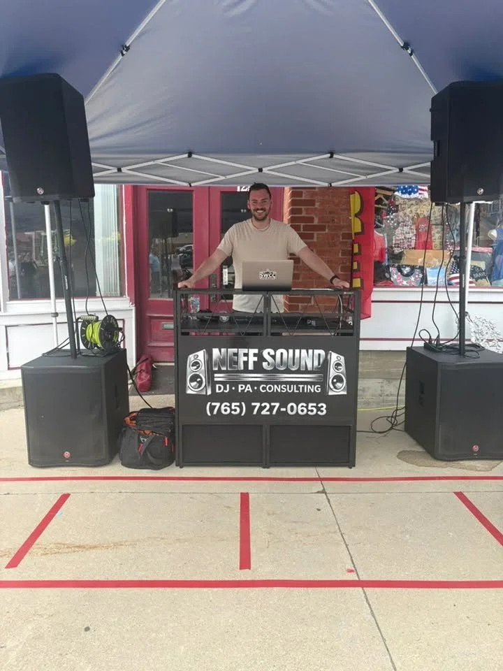 DJ standing behind sound equipment booth with large speakers and signage for Neff Sound, DJ PA consulting, at outdoor event