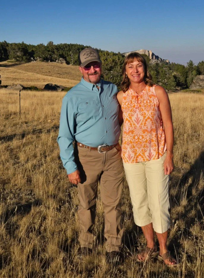 A man and a woman standing outdoors in a grassy field, smiling at the camera. The man is wearing a light blue shirt, khaki pants, and a baseball cap. The woman is wearing an orange patterned sleeveless top and white capri pants. There are trees and a hill or rock formation in the background.