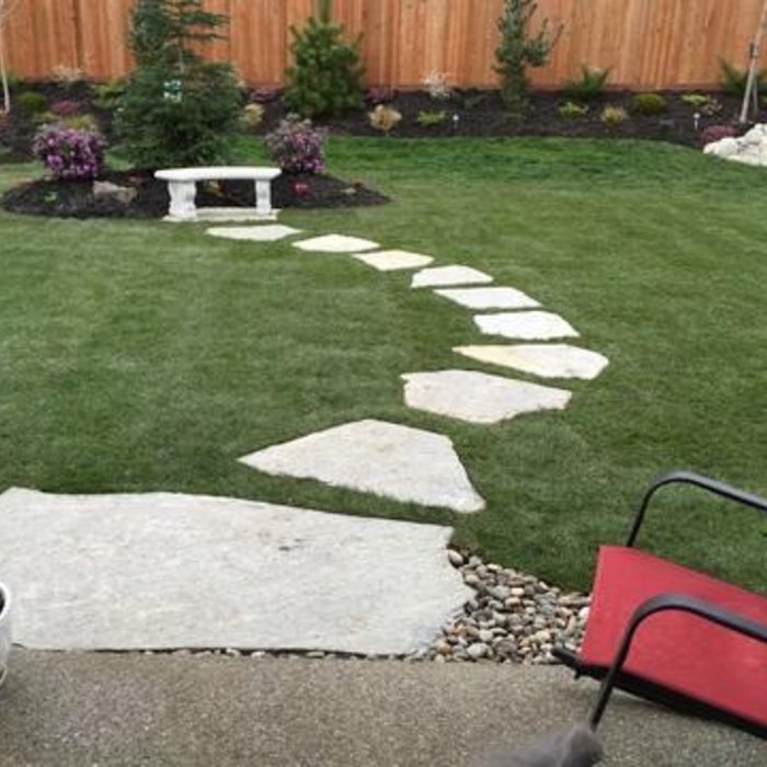 Rocky Creek Landscape & Design sample of work in Boise Idaho. Curved stone pathway with grass and garden bed in backyard, including a white bench and small trees, with a red chair and part of a patio in the foreground.