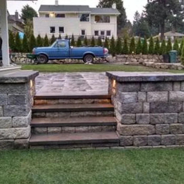 A stone staircase with three steps leads up to a paved patio area, with a stone wall on either side. In the background, there is a blue vintage pickup truck parked on the grass and a house with a white exterior and multiple windows. There are trees and a trash bin in the yard.