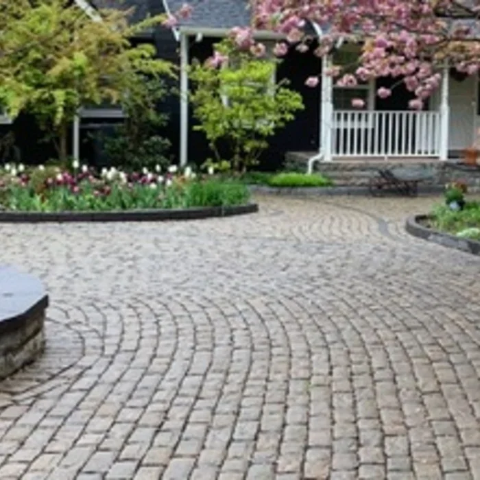 Brick driveway in Boise Idaho leading to a porch surrounded by colorful blooming flowers and green trees, with a white railing and pink flowering tree in the background.
