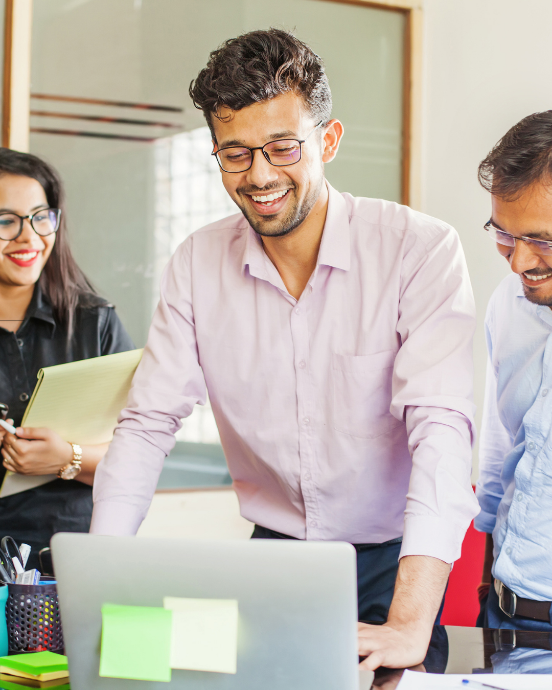 A group of diverse coworkers in a meeting, smiling and working together in an office, with a laptop and sticky notes on the table.
