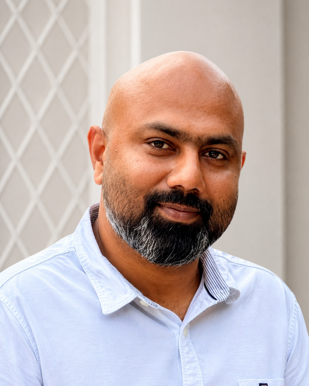 A close-up portrait of a man with a bald head, dark beard with some gray, wearing a light blue collared shirt, standing outdoors with a neutral background.