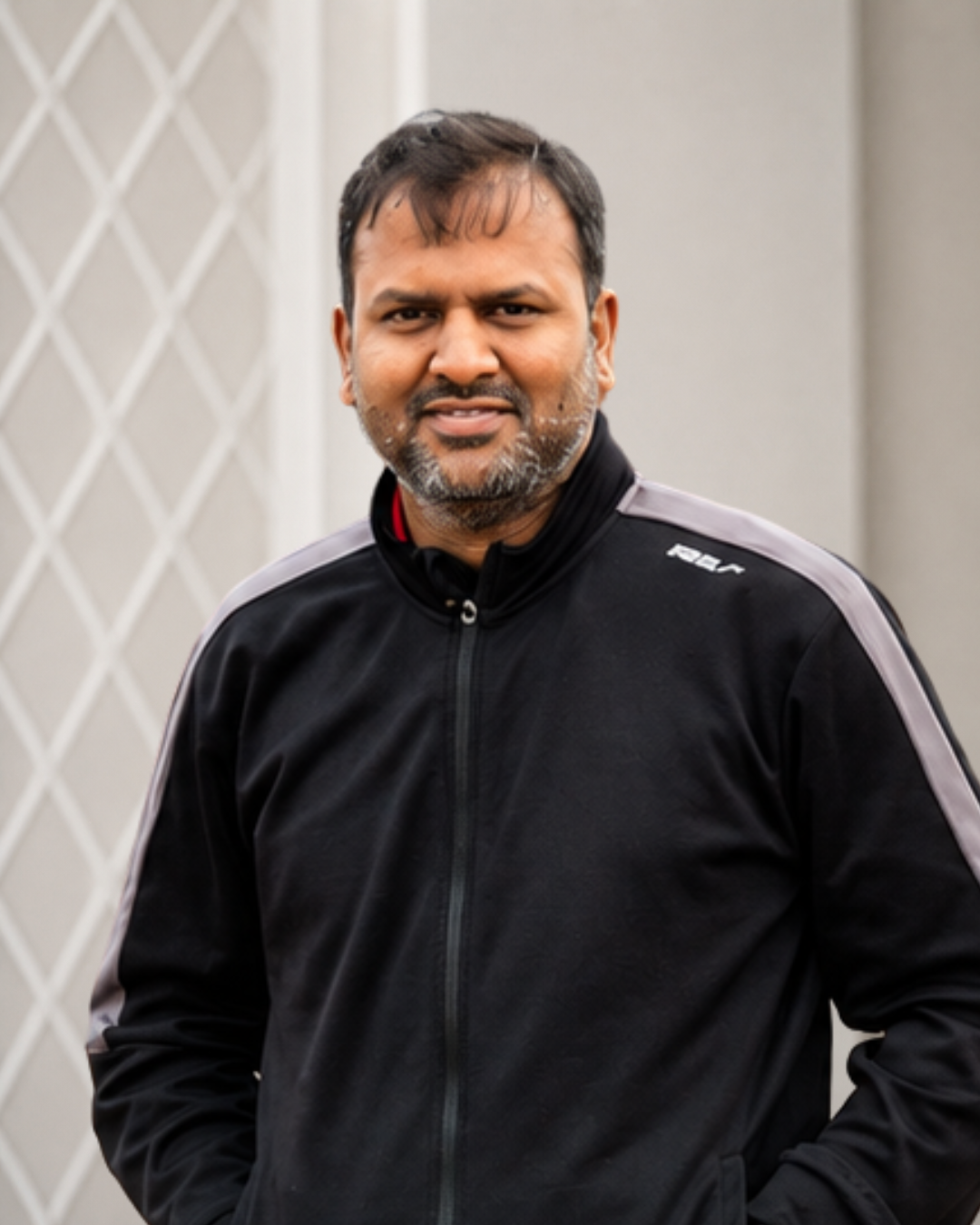 Man with dark hair and beard wearing a black track jacket with gray accents, standing outdoors in front of a white lattice-patterned wall.
