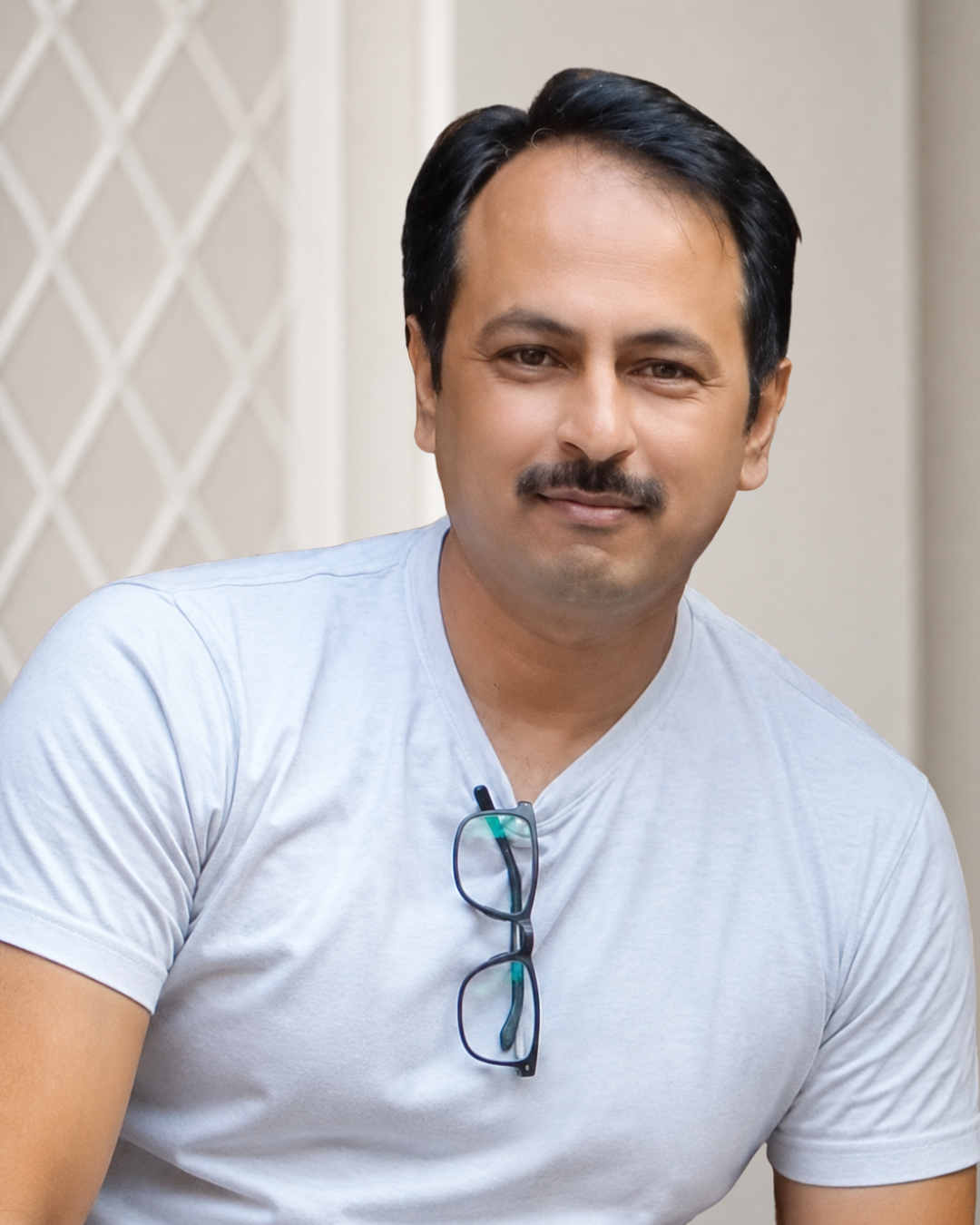 A man with dark hair, a mustache, wearing a light gray t-shirt and glasses hanging from his shirt collar, smiling softly at the camera.