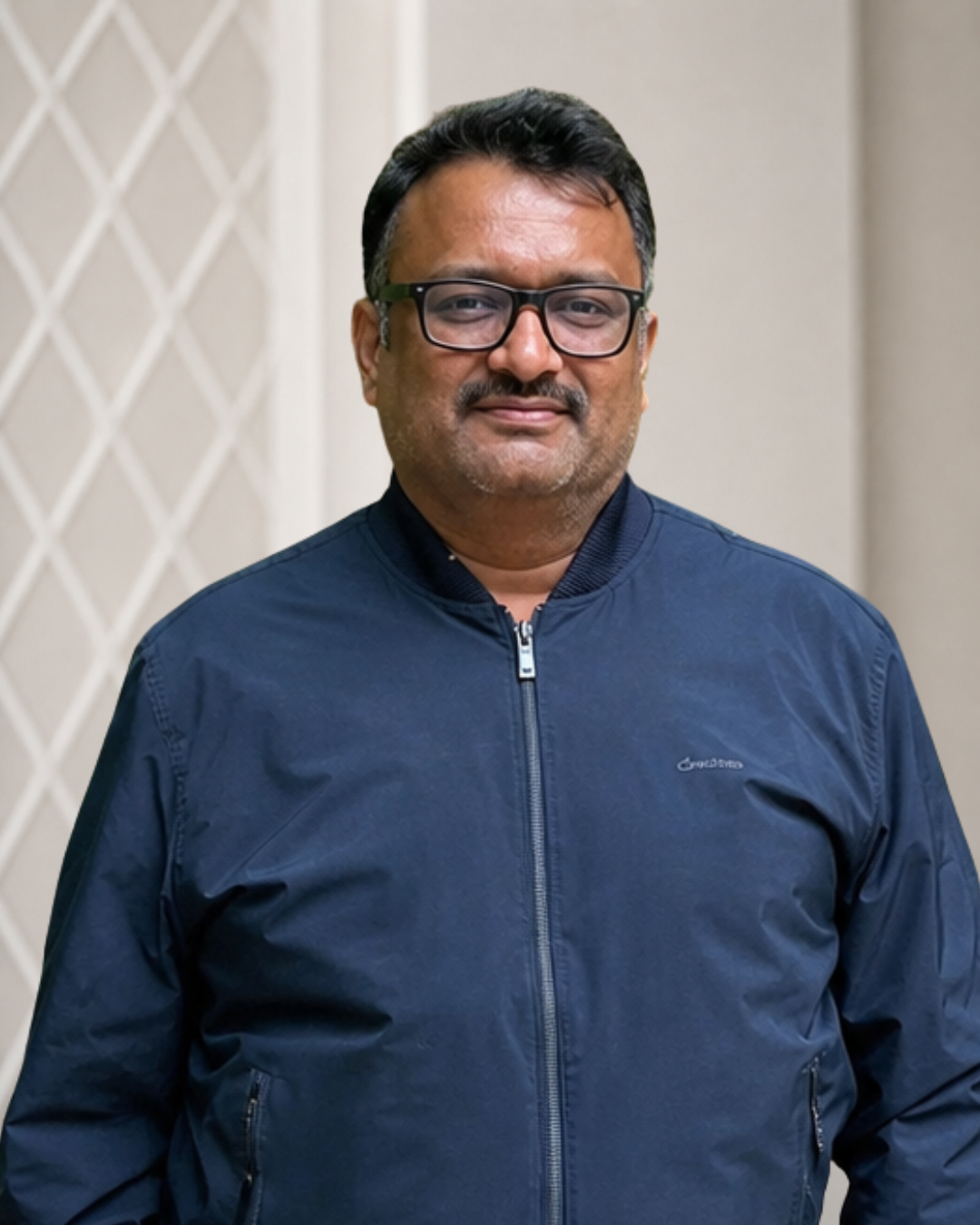 A man wearing glasses and a blue jacket with a neutral background.