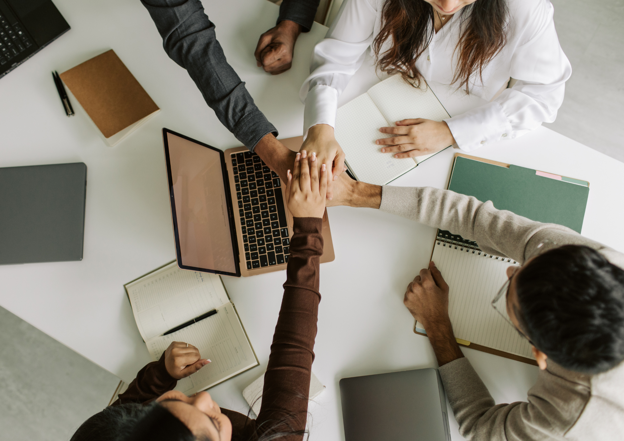 Five people sitting around a white table with laptops, notebooks, and pens, holding hands in the center for a group handshake or gesture.