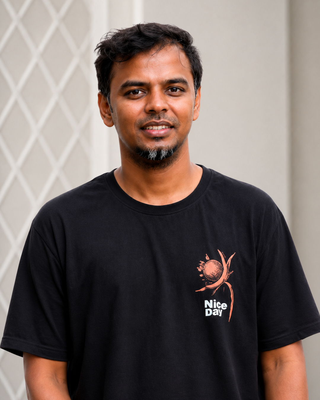 A man with short black hair, a goatee, and a slight smile, wearing a black T-shirt with a logo and text that says 'Nice Day,' standing outdoors in front of a wall with a lattice design.