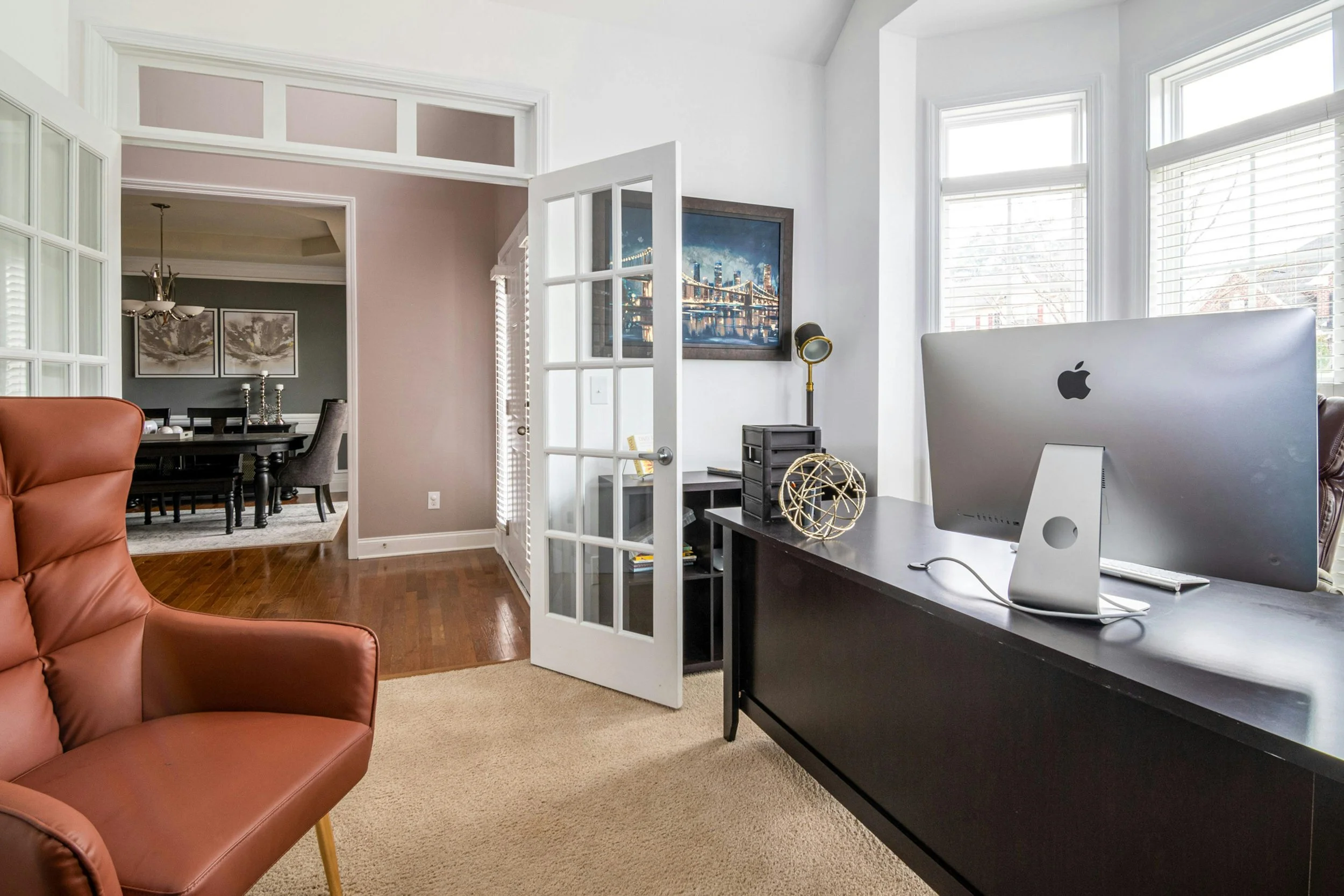 A home office with a black desk, Apple iMac computer, decorative globe, and a black file organizer. Large windows with white blinds, and a view of neighboring houses outside. Part of a dining area with a dark dining table and chairs is visible in the background.