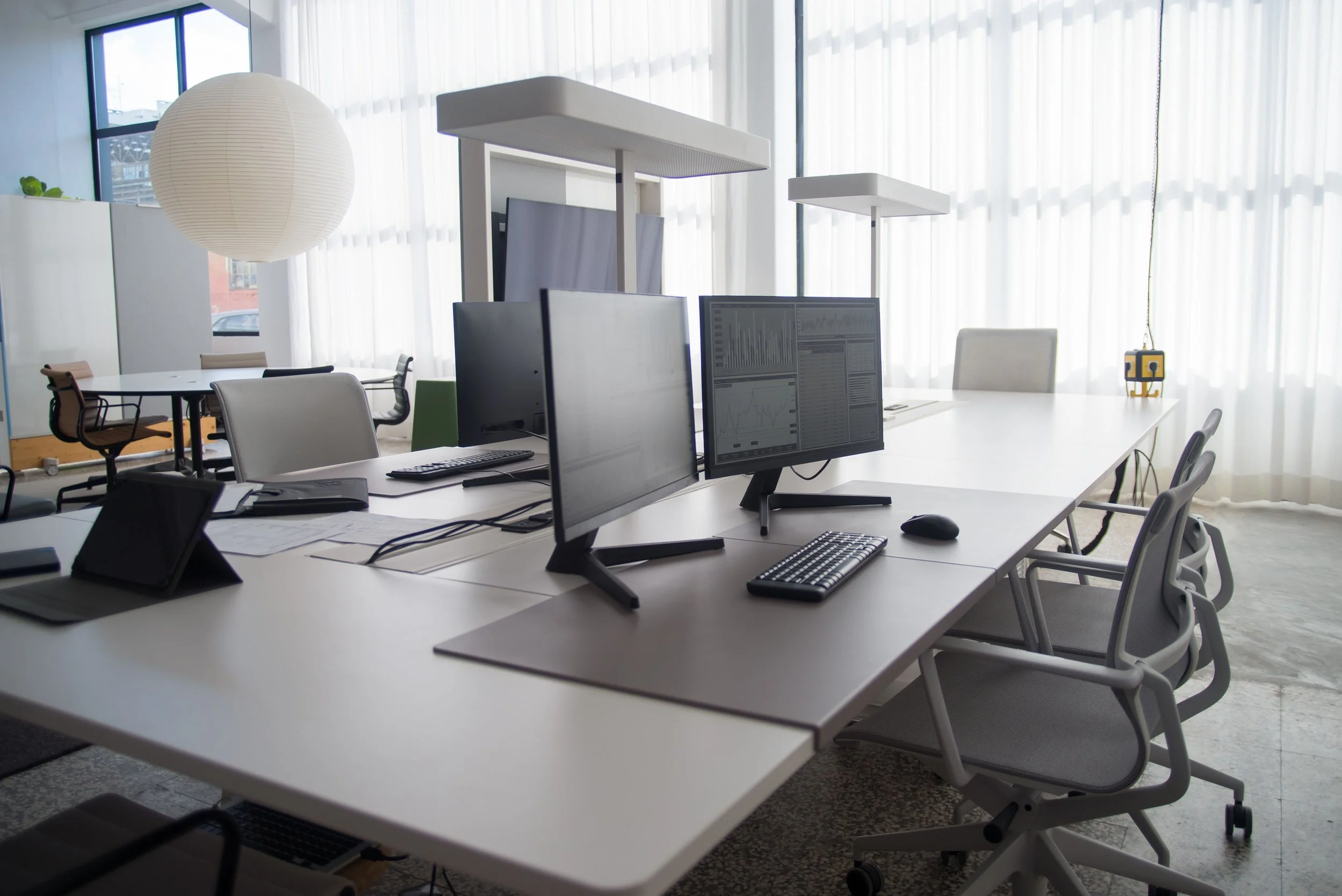 Modern office with multiple white desks, computer monitors, chairs, large windows with sheer white curtains, and some office supplies.