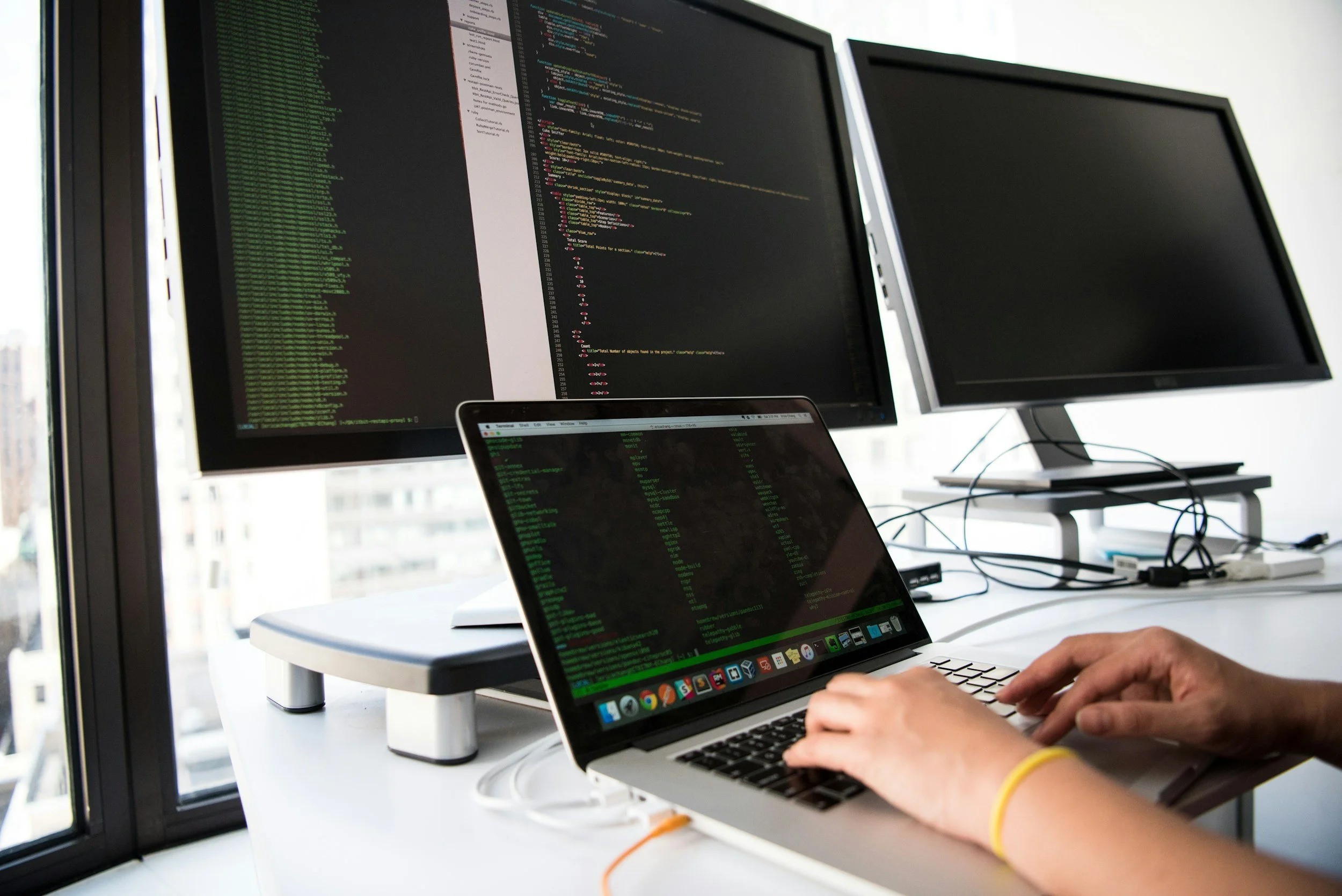 A person working on a laptop with code displayed on the screen, with two large monitors in the background also showing code, set in a bright office near large windows.