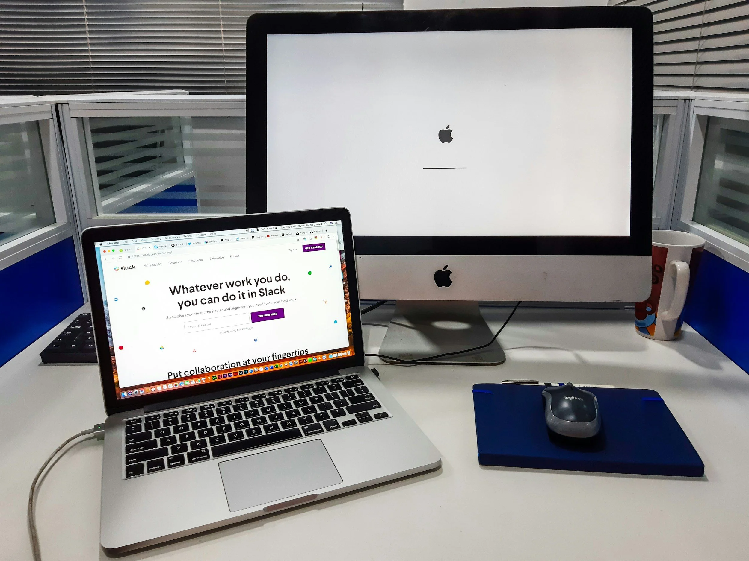 A workspace with a MacBook laptop displaying Slack's homepage, an Apple desktop monitor showing an Apple logo and loading bar, a Logitech mouse on a blue mouse pad, a mug with colorful design, and a white desk with partitioned glass panels in the background.