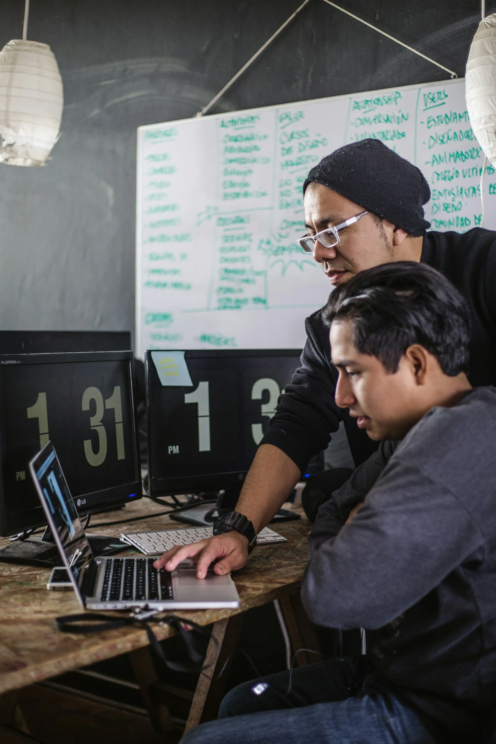 Two young men working on laptops at a rustic wooden table in a tech workspace, with computer monitors displaying the number 13,31, and 1, containing a whiteboard with handwritten notes and a chalkboard wall in the background.