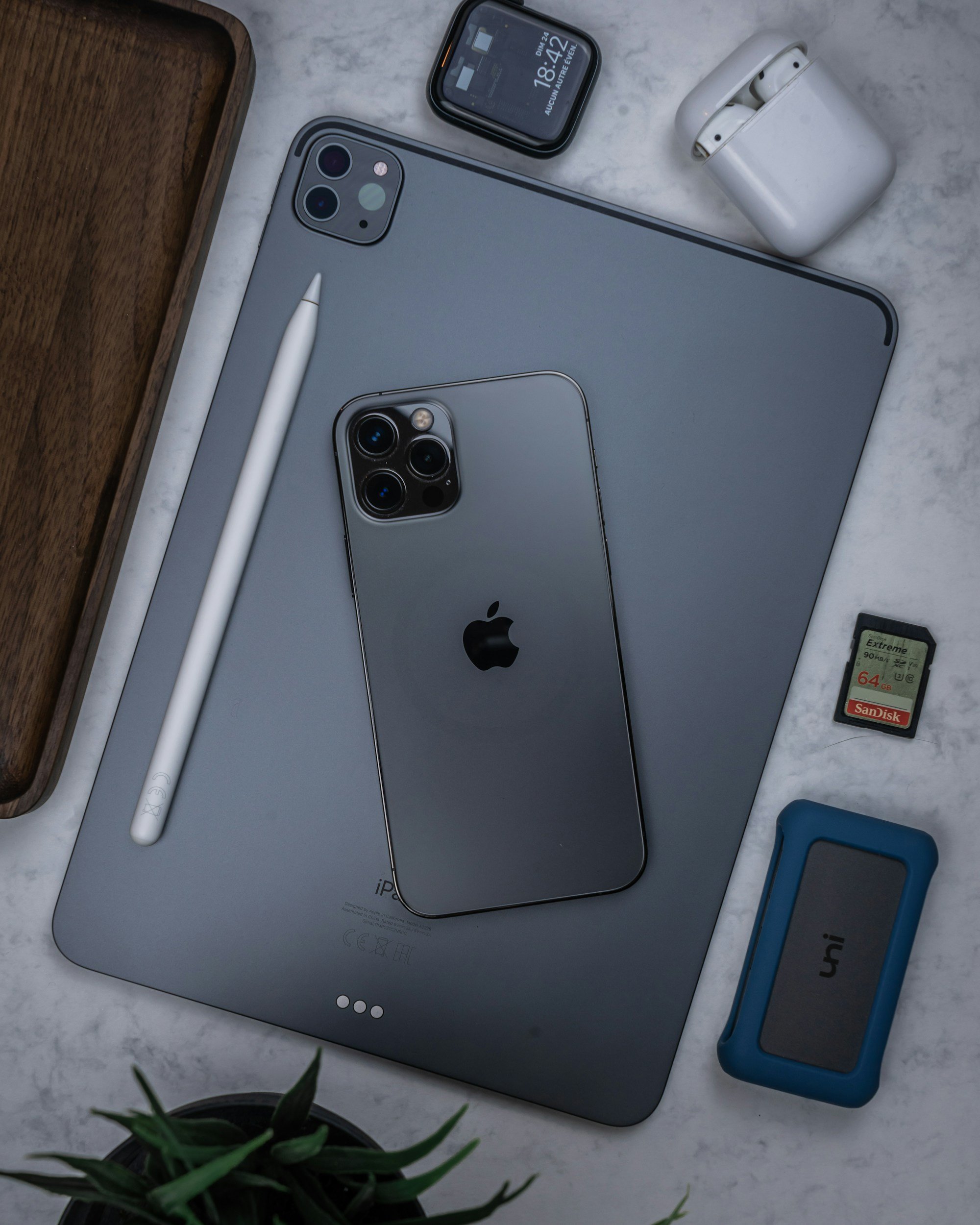 An iPad, iPhone, Apple Pencil, wireless earbuds with case, microSD card, and a blue Apple Watch band arranged on a white marble surface, with a small potted plant in the foreground.