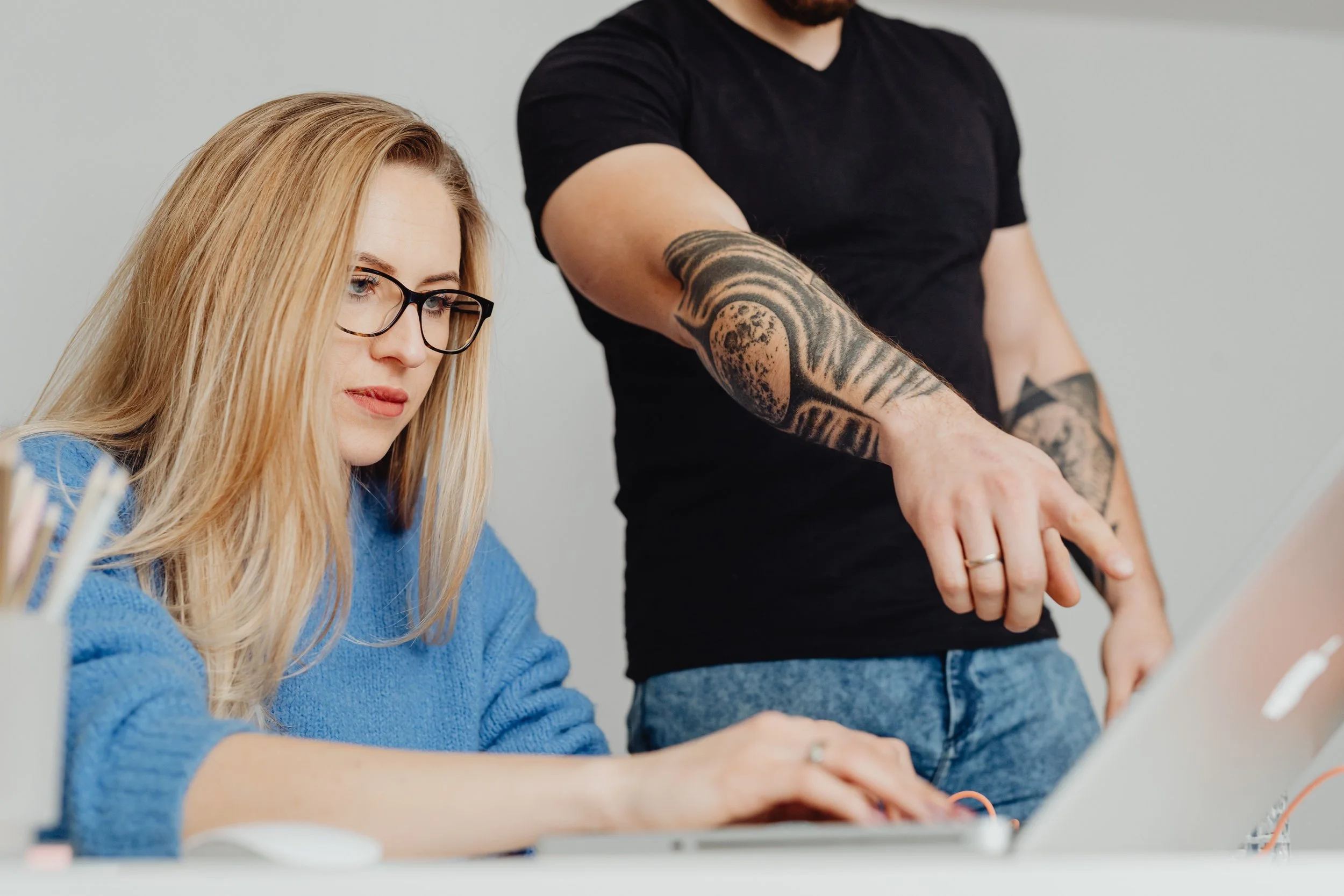 A woman with blonde hair and glasses working at a desk with another person showing their tattooed arm.