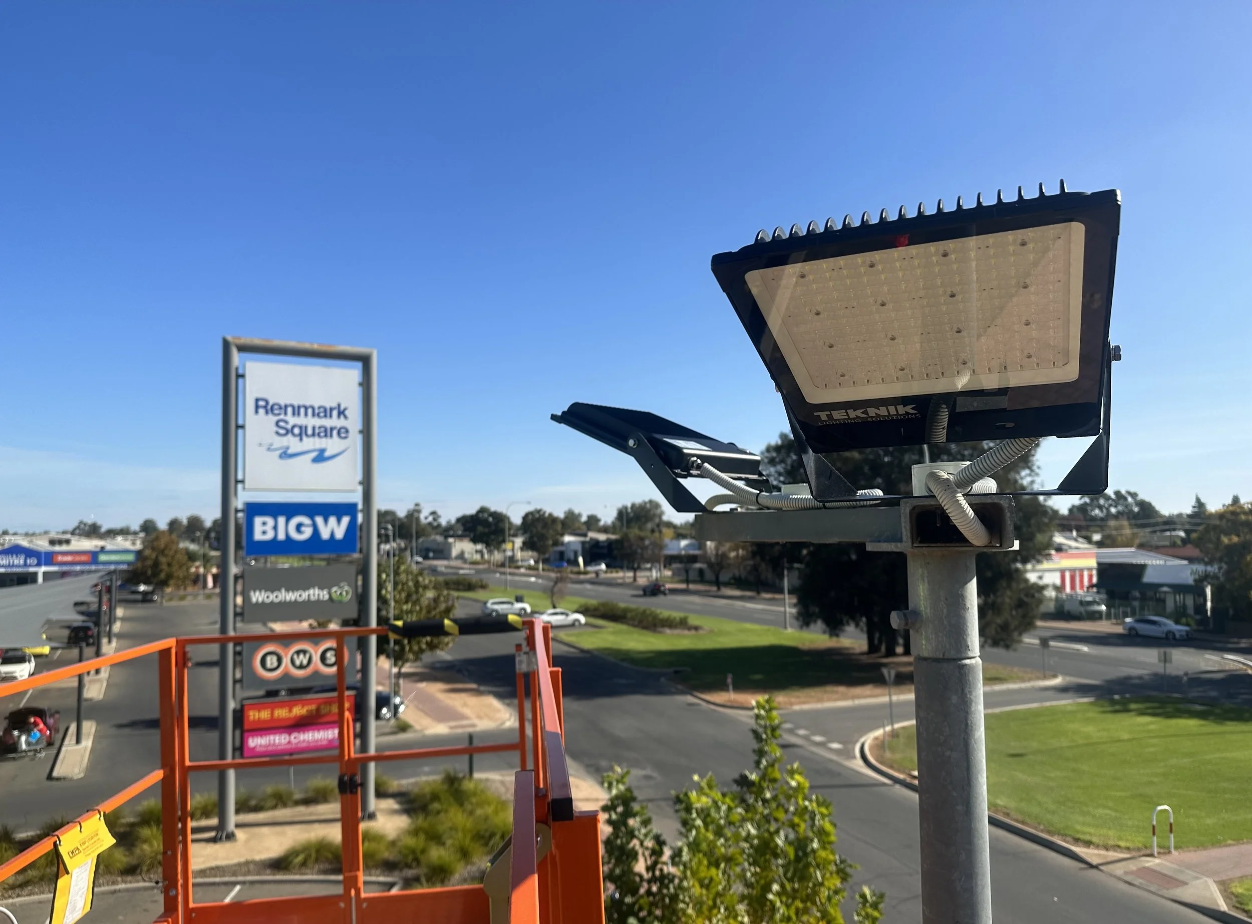 Close-up view of a solar LED street light mounted on a pole, with a shopping center sign for 'Benmark Square' and other commercial signs visible in the background on a sunny day.
