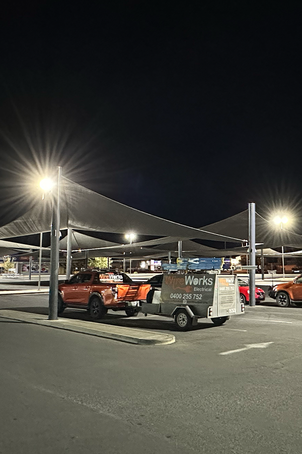 A parking lot at night with a canopy structure, lit by bright spotlights, with a red pickup truck and a utility trailer labeled 'Wire Works Electrical'.