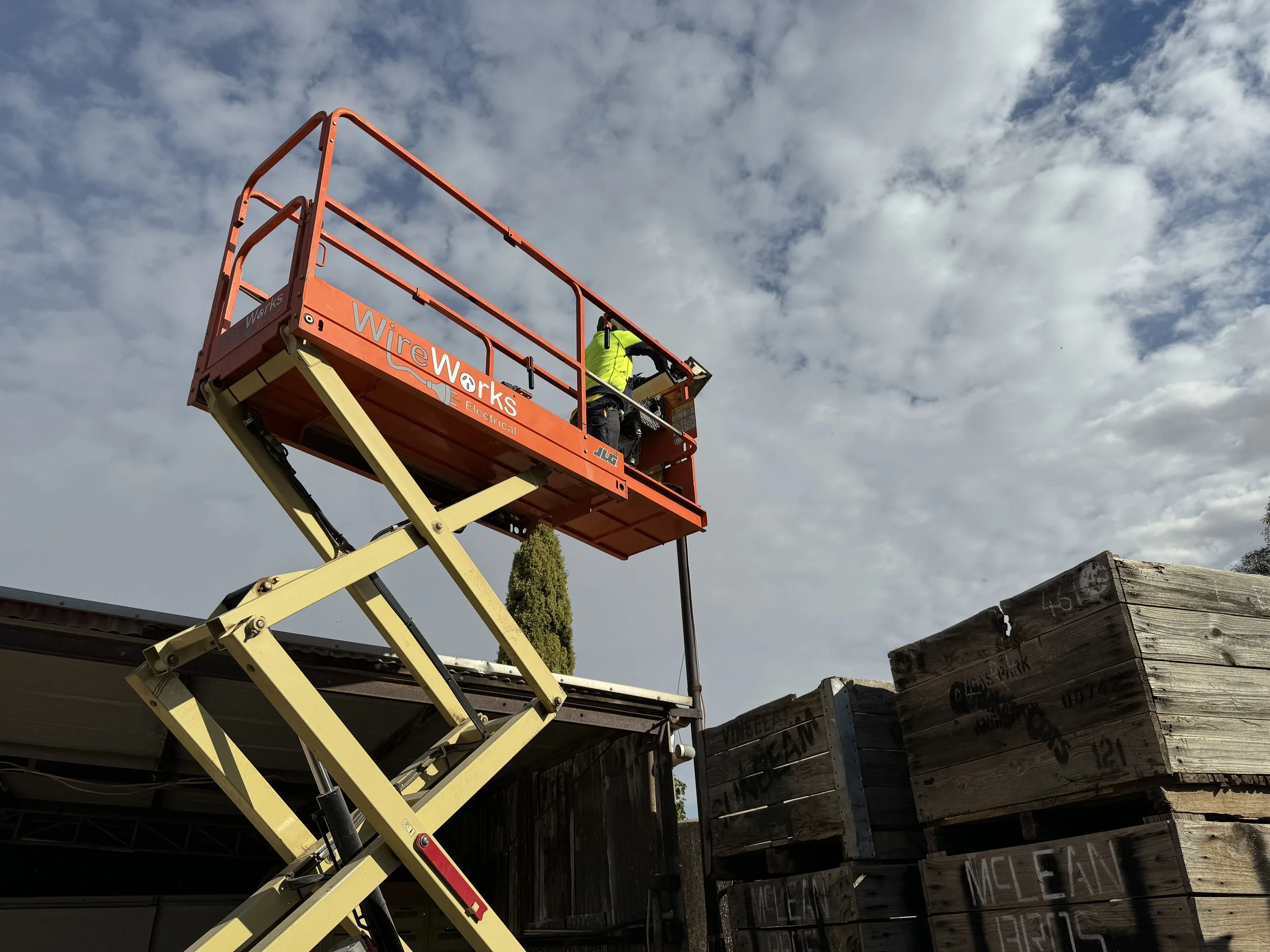 A worker in a yellow safety vest on a cherry picker lifts a pole or wire against a cloudy sky.