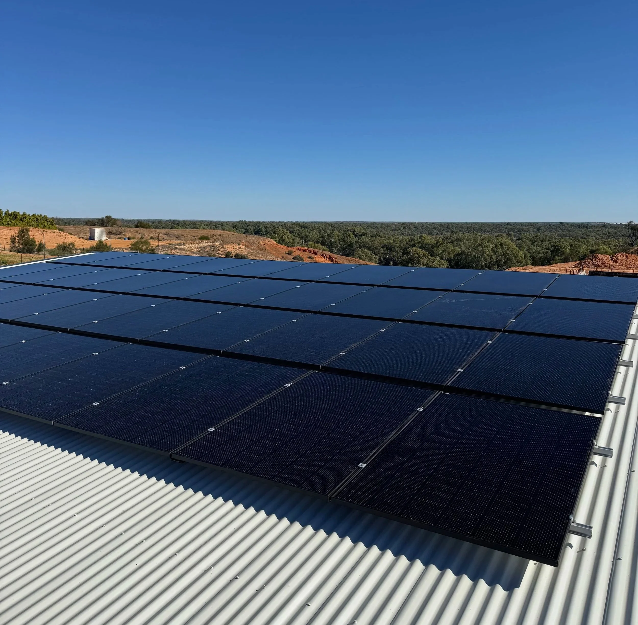 A landscape with a clear blue sky, green trees, and a solar panel installation on a white corrugated metal roof.
