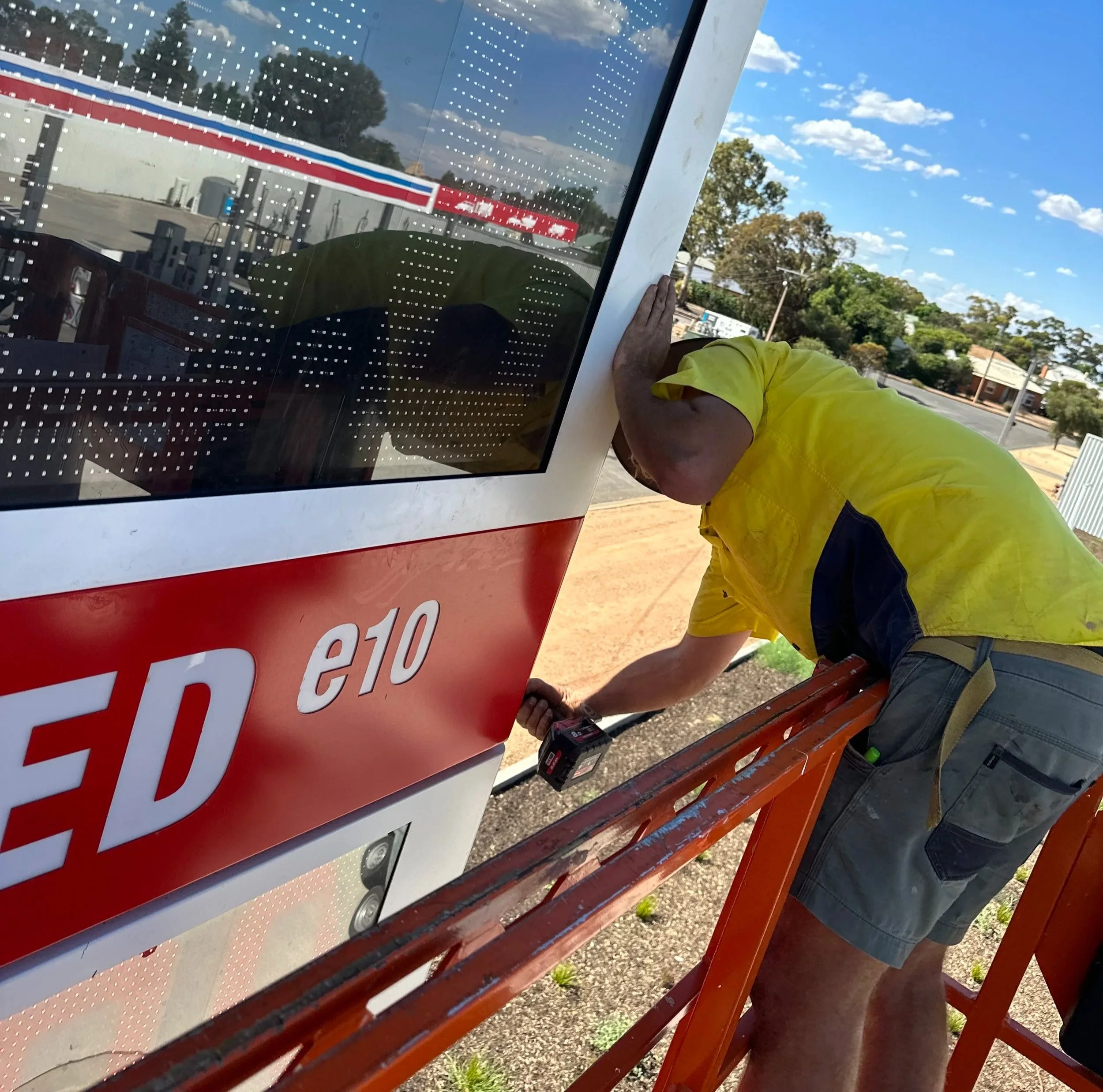 A person in a yellow shirt and shorts is working on an outdoor signboard, with a power drill in hand, next to a red wooden bench on a sunny day with trees and clouds visible in the background.