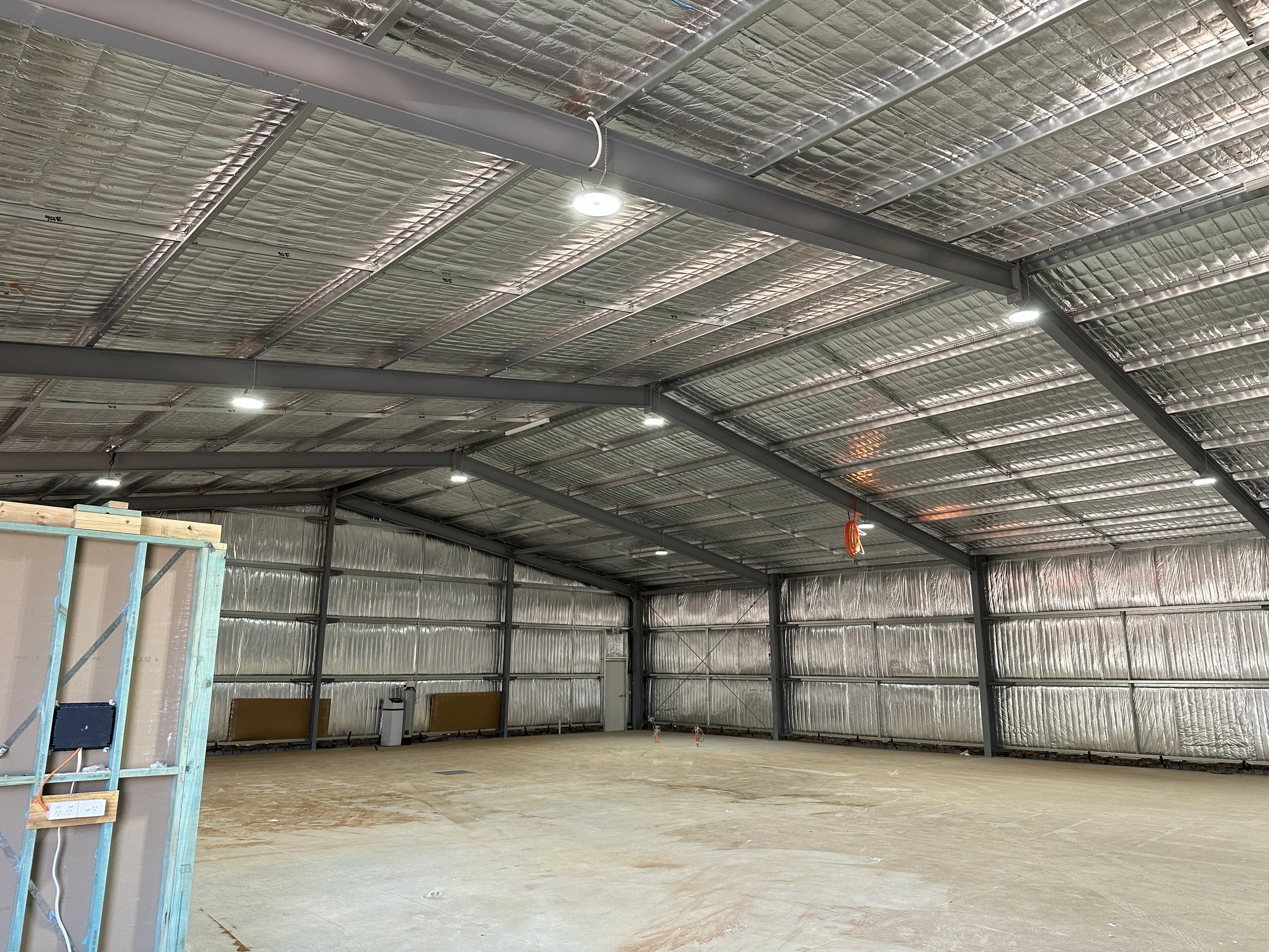 Interior of a building under construction, showing exposed metal roof beams and insulation, with a partially finished wall and construction materials.