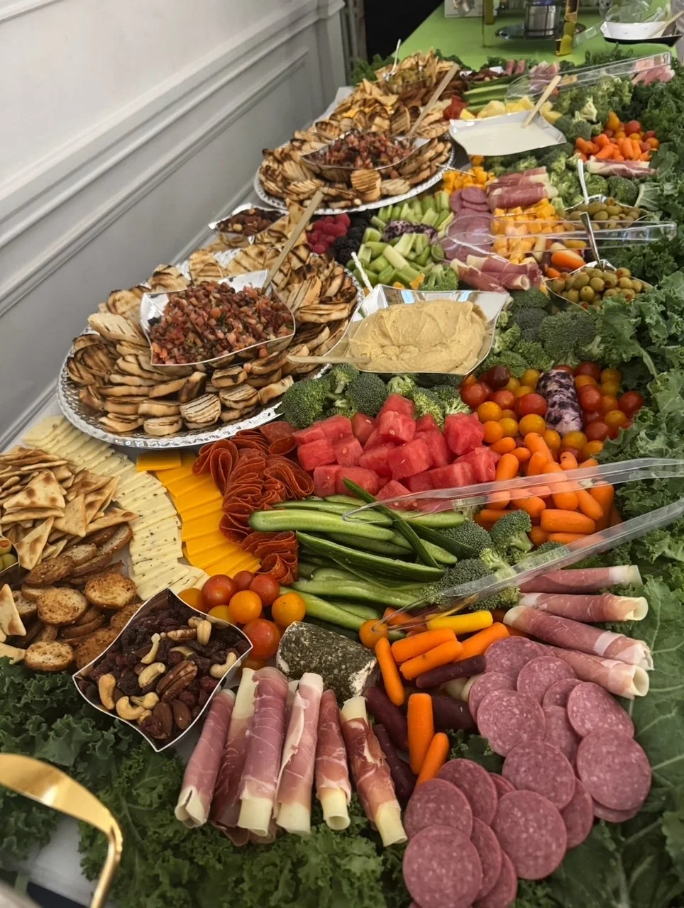A long table filled with various appetizers including vegetables, sliced meats, cheeses, nuts, fruits, and crackers, arranged for a party or gathering.