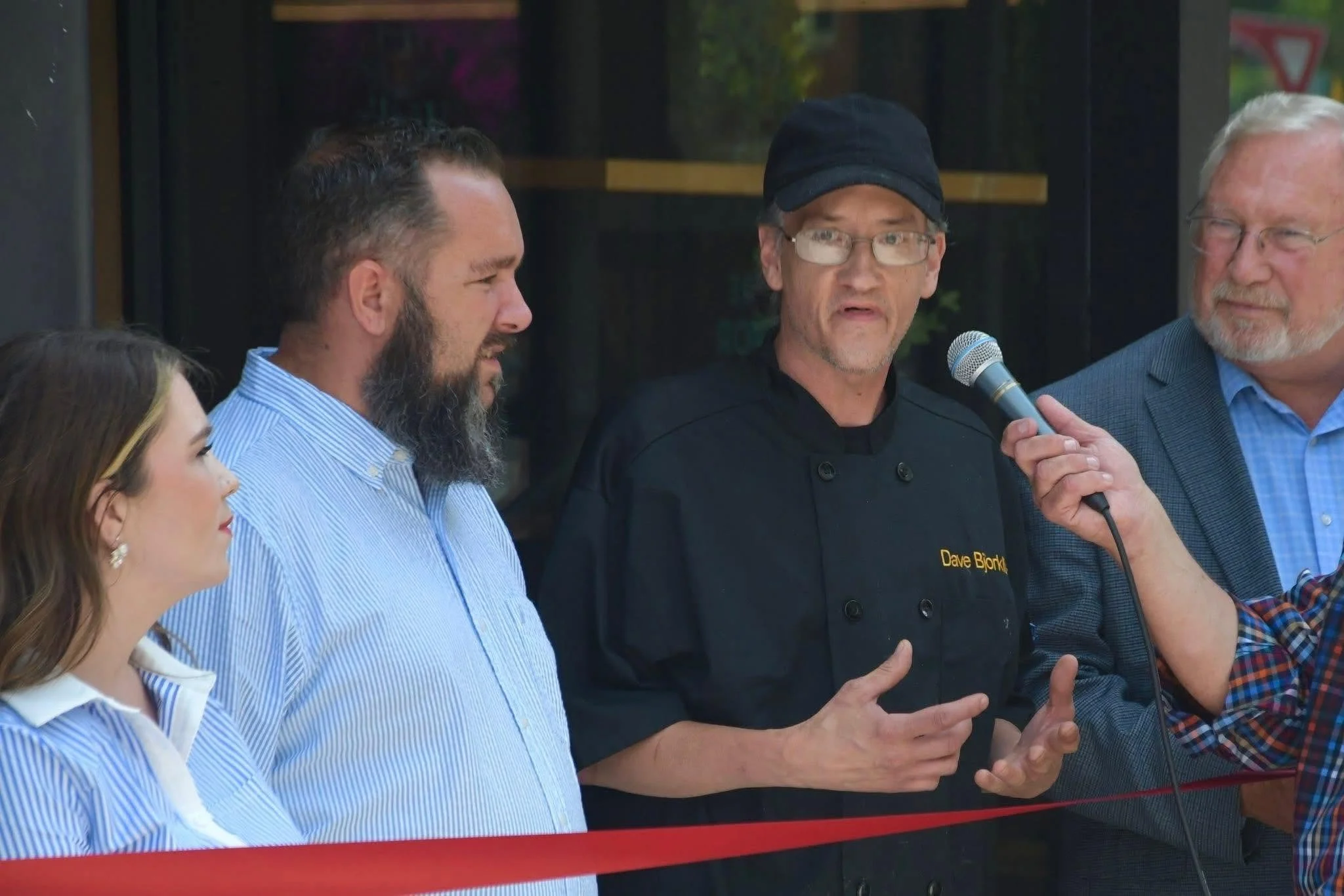 A group of four people standing together outdoors, with one person speaking into a microphone. The man with glasses and a black cap wearing a black chef's jacket labeled 'Dave Bjork' appears to be the focus of attention. The other individuals, including a woman and two men, are listening closely.