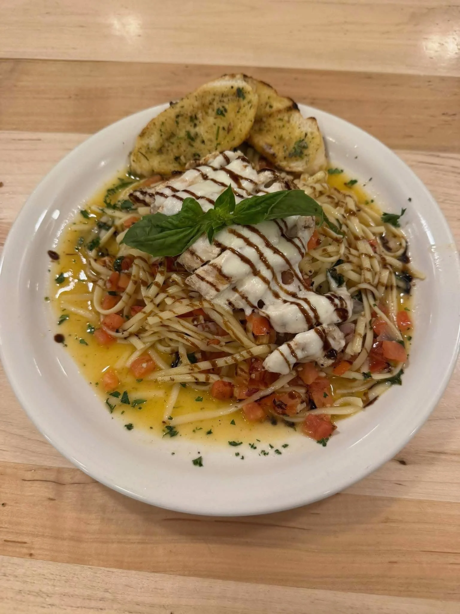 A white plate of pasta with grilled chicken topped with basil and balsamic glaze, served with grilled bread on a wooden table.