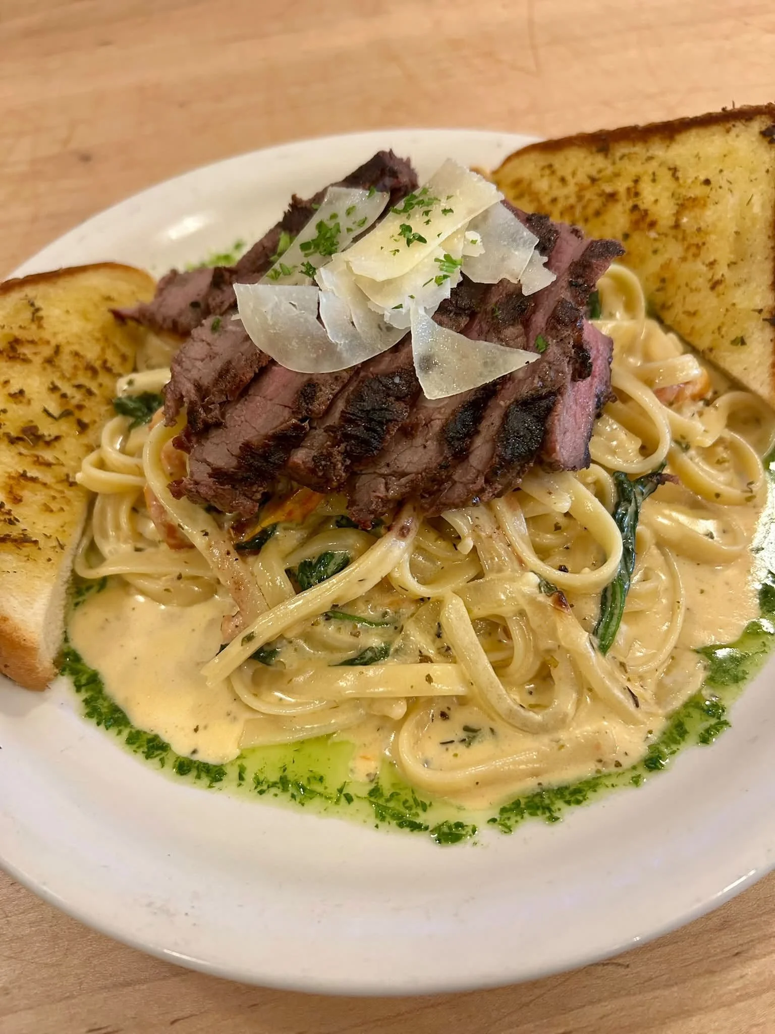 A bowl of creamy pasta topped with slices of grilled steak, shaved cheese, and garnished with chopped herbs, served with toasted garlic bread on the side.