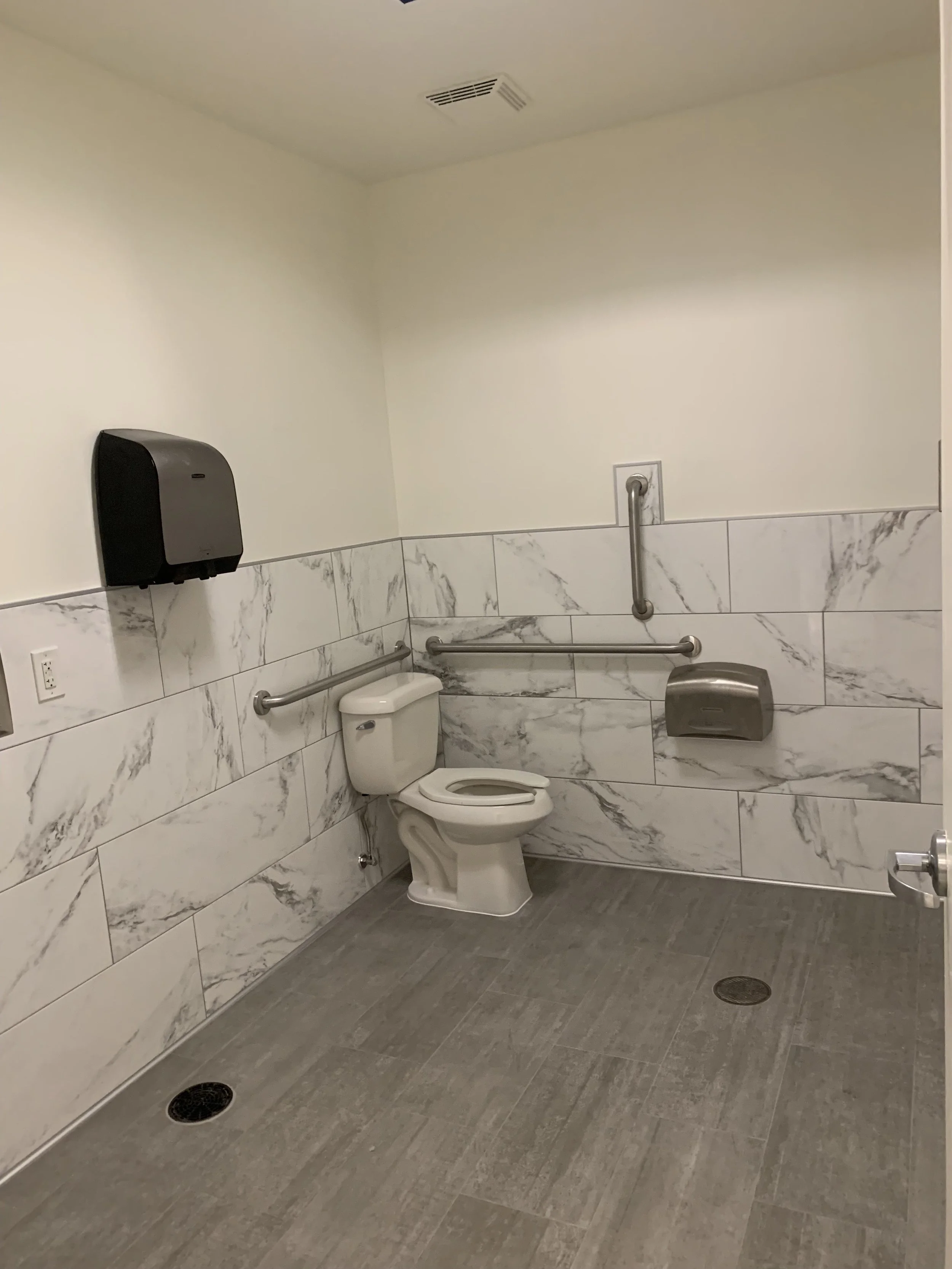 Accessible bathroom with a toilet, safety grab bars, soap dispenser, and a paper towel dispenser on the wall.
