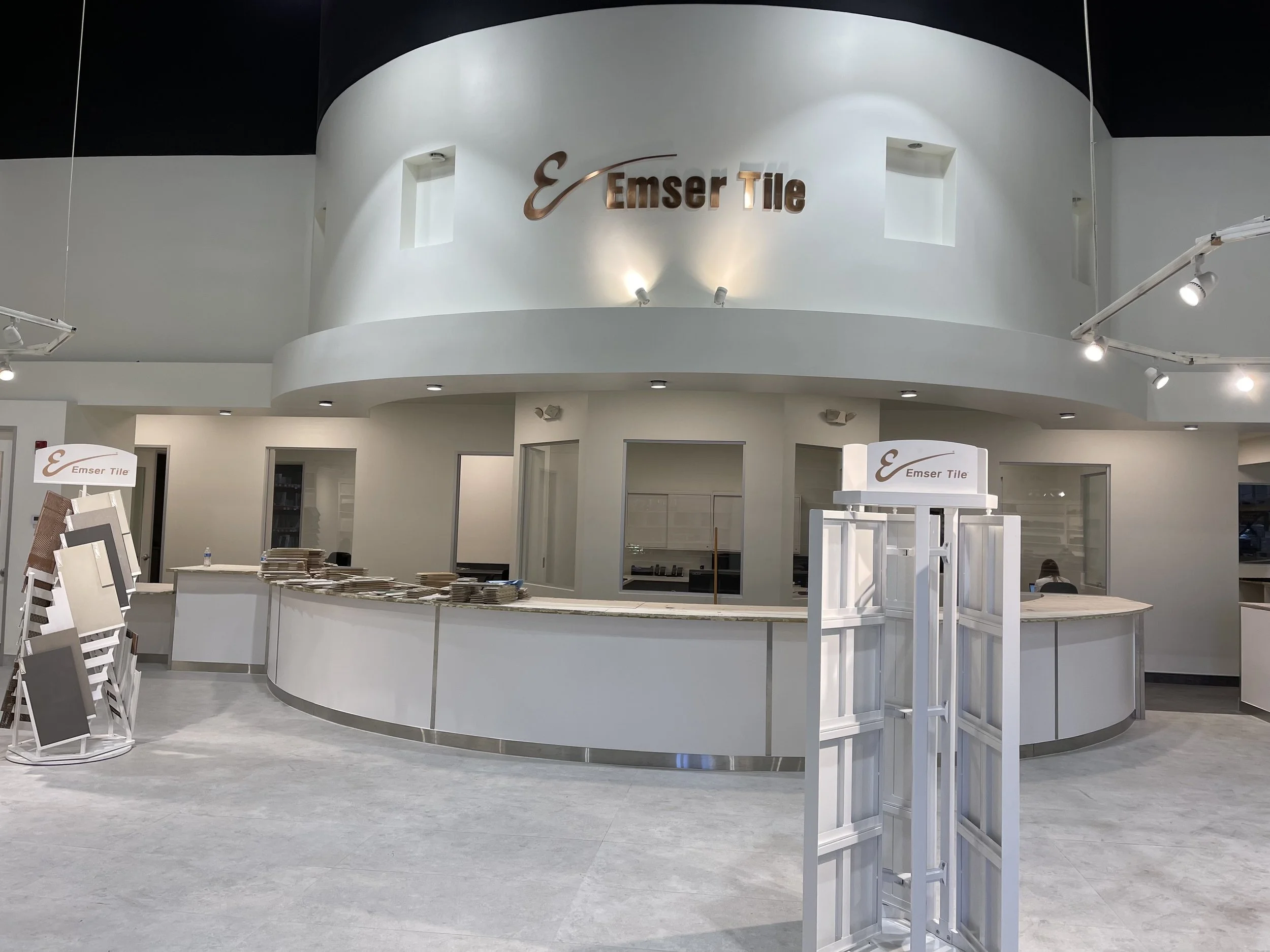 Interior of a showroom with a curved white reception counter displaying samples of tiles, a prominent sign reading 'Emser Tile' on the wall above, and various display racks and shelves around the space.