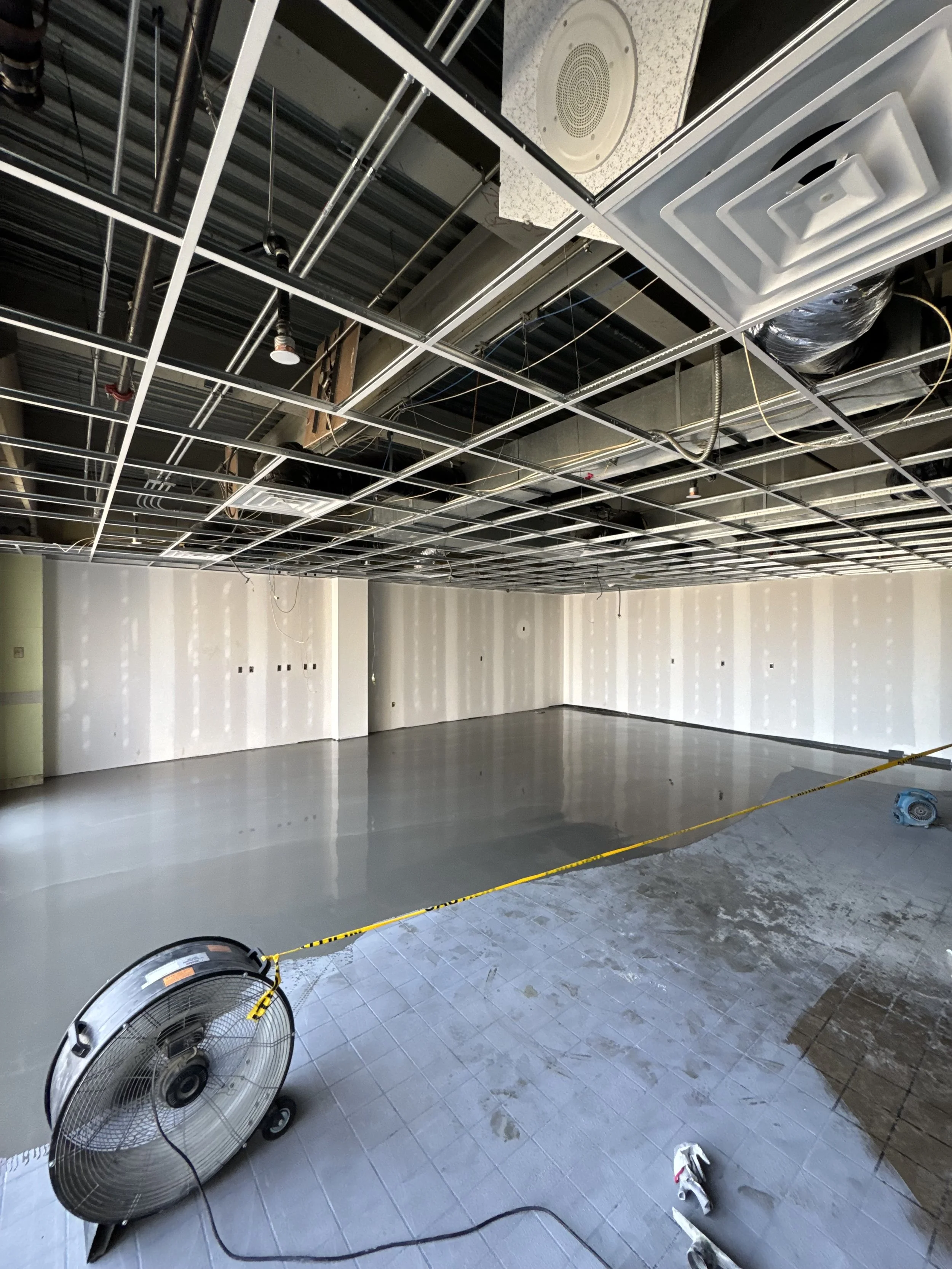 Construction site with unfinished ceiling revealing metal framework, ductwork, and pipes, drywall on walls, and construction tools and equipment on the floor.
