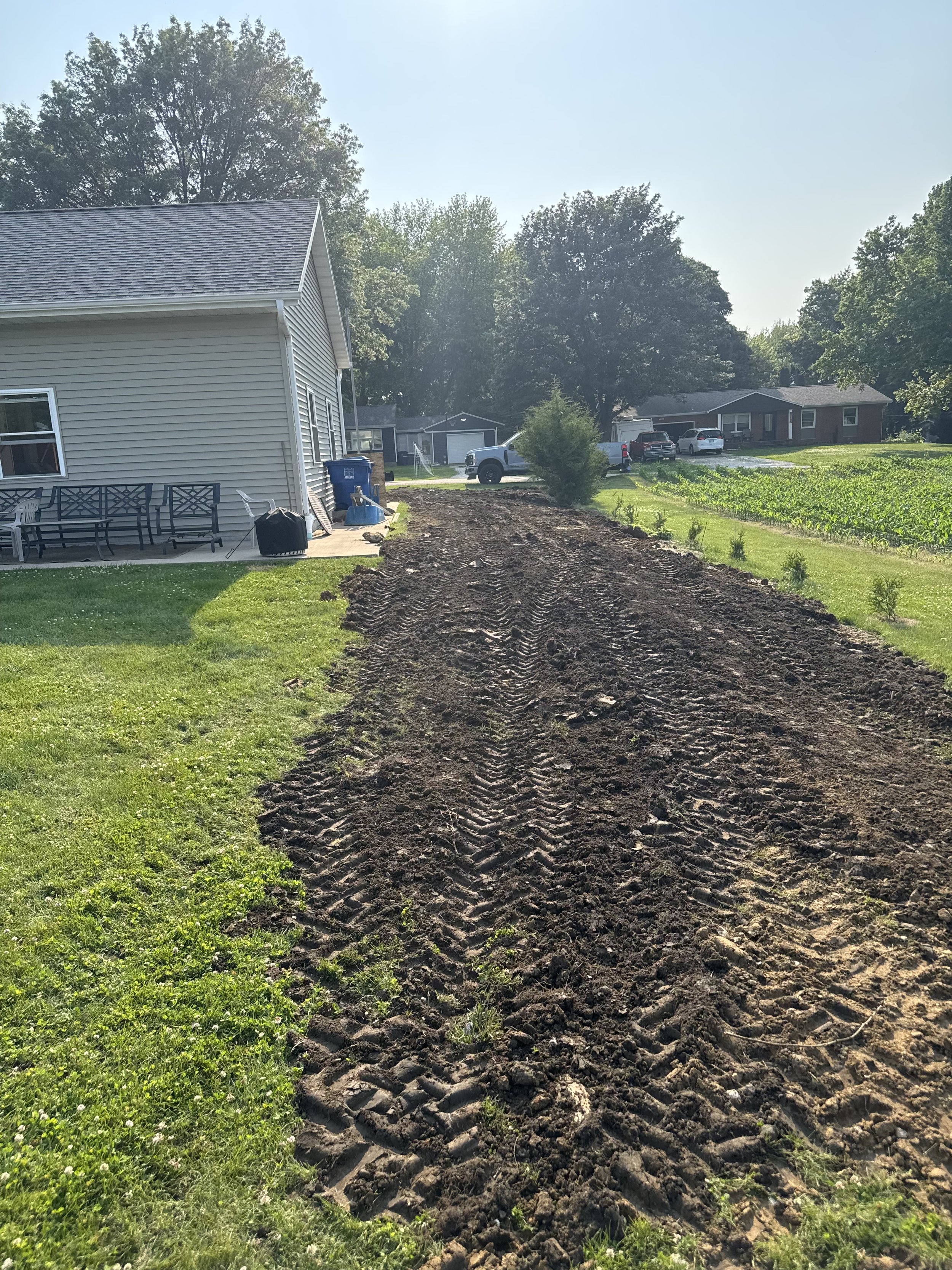 A backyard with freshly tilled soil in a straight line, adjacent to green grass, with a beige house on the left, and trees and parked cars in the background on a sunny day.