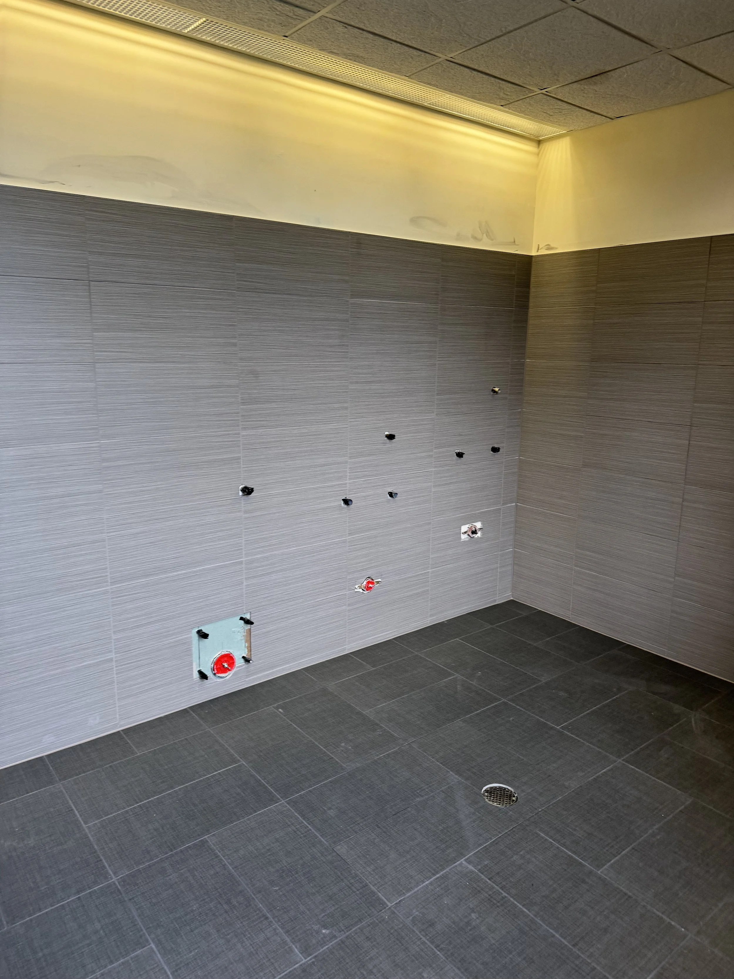 A partially completed bathroom or washroom with gray tiled walls and floor, with visible unfinished electrical outlets and wiring, and a drain on the floor.