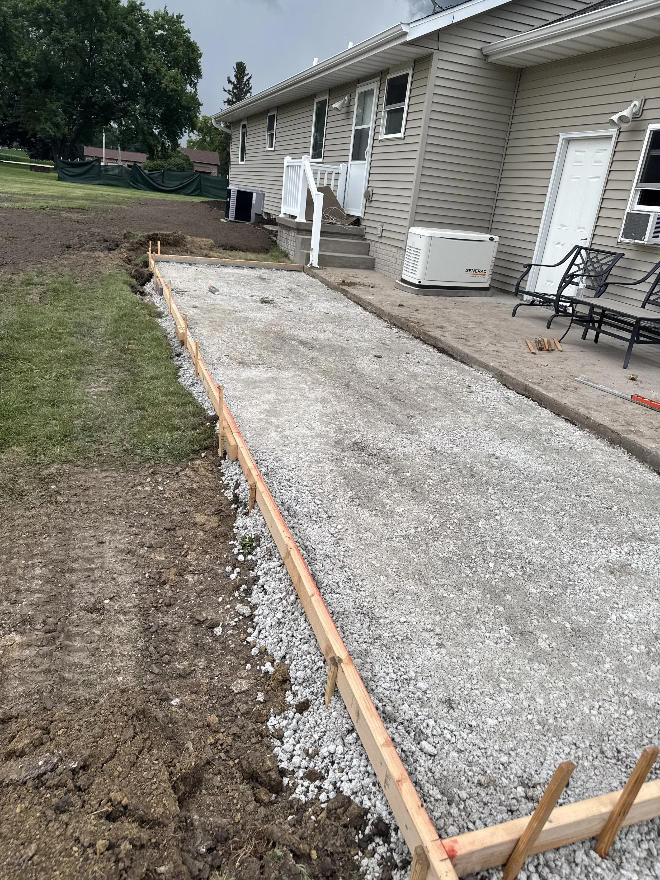 A backyard patio area under construction with gravel, wooden framing, and a row of benches. The house has beige siding, and there are smoke detectors and air conditioning units visible. The area appears to be prepared for paving or concrete work.