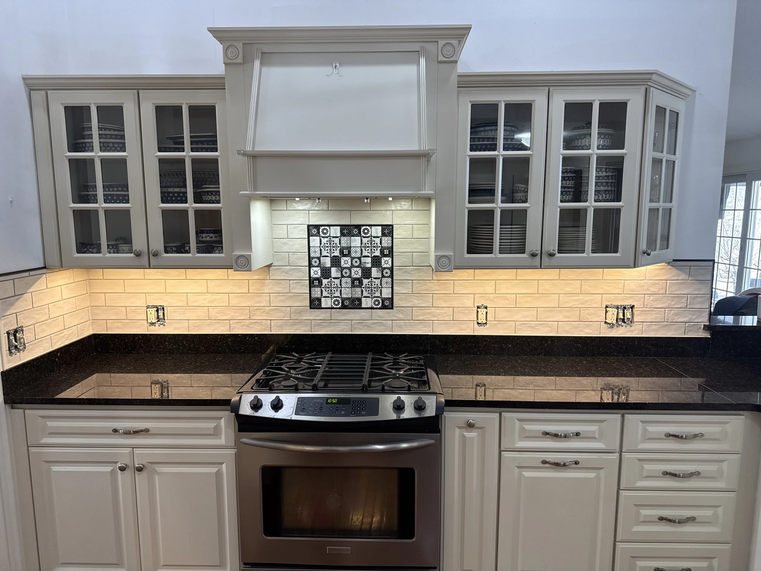 White kitchen cabinets with glass doors, a stove, and a beige tile backsplash. There is a black stove top with an oven below, and unconnected electrical outlets on the wall.