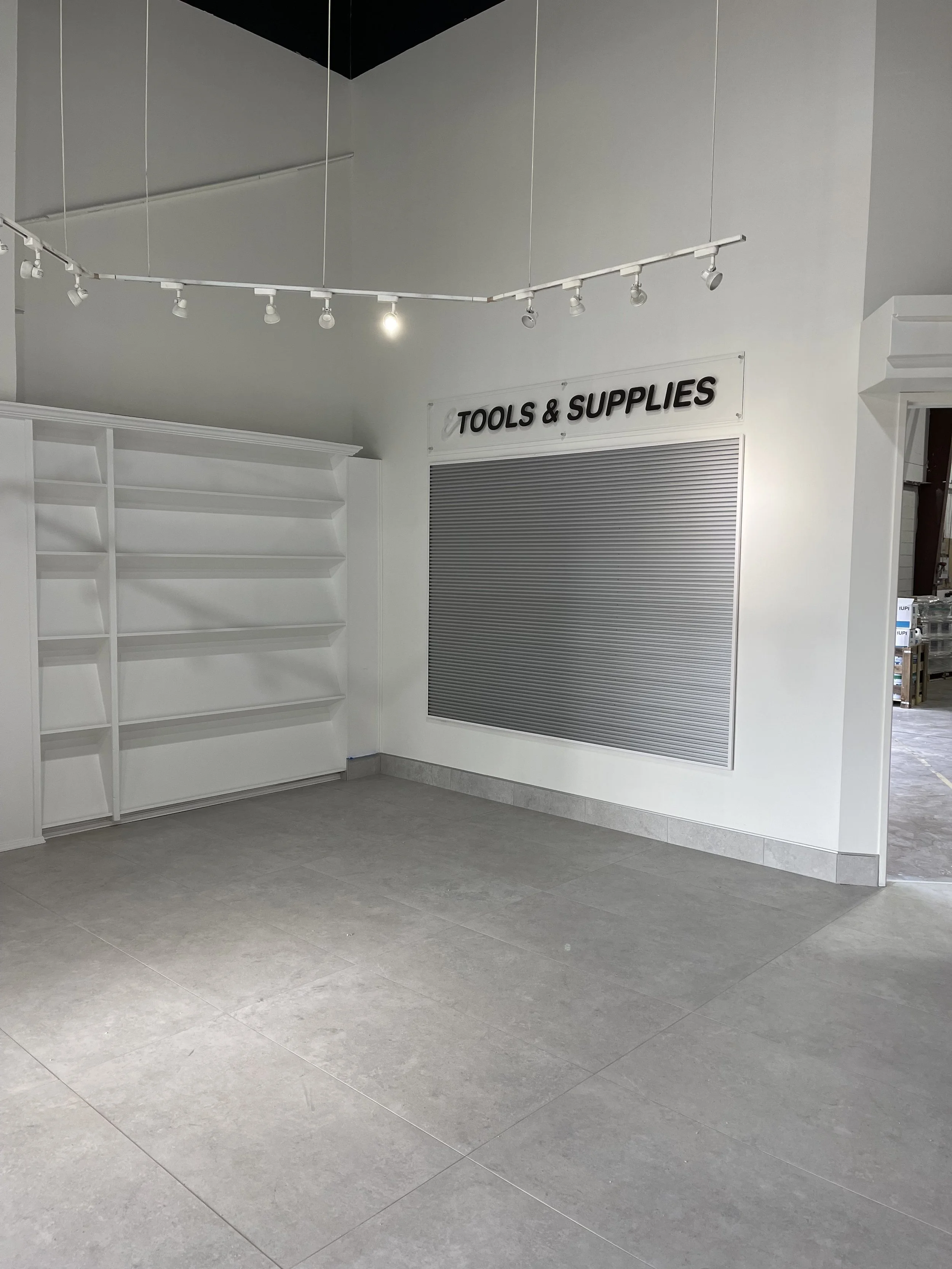 Empty store section with white shelves, closed tool and supplies shutters, and gray tiled floor.