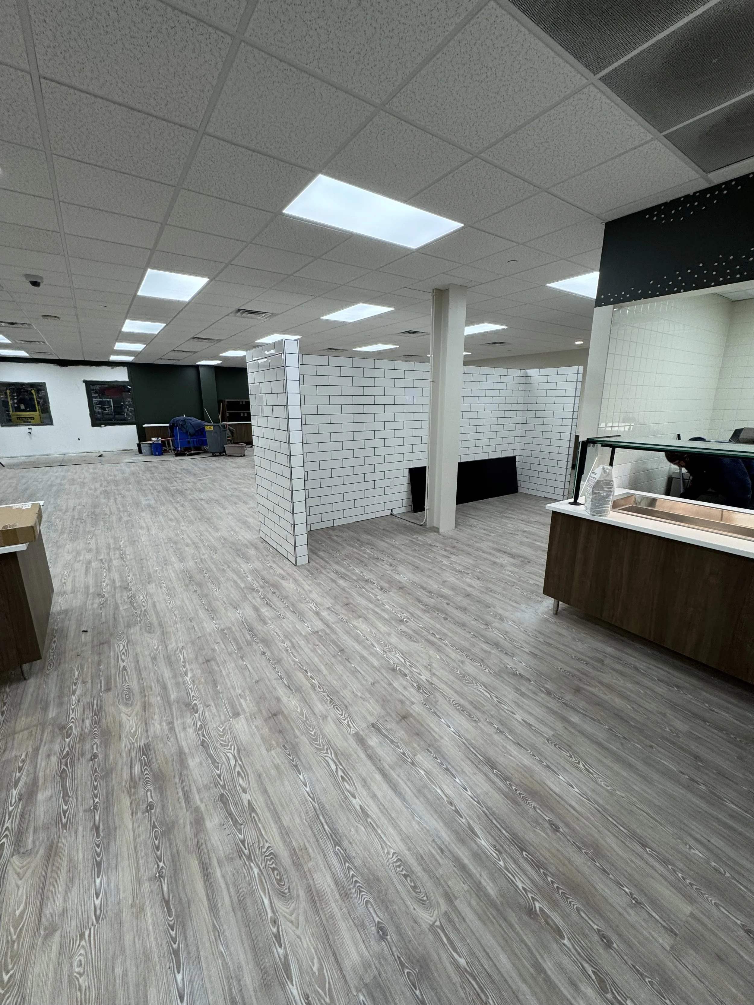 Interior view of a commercial space under construction or renovation, featuring drop ceiling lights, white tiled partial walls, a wood floor, and some construction equipment and supplies.