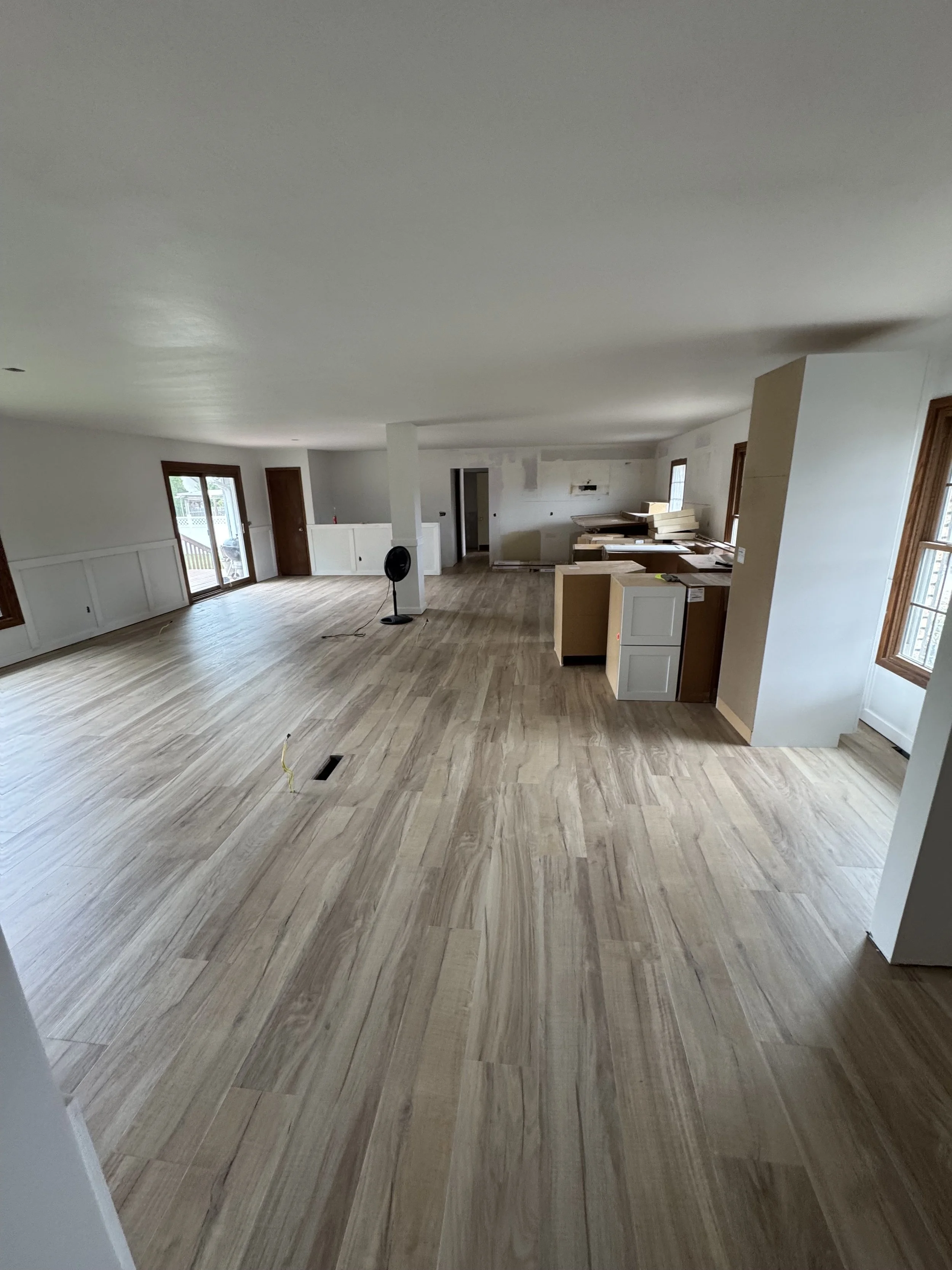 A living room under construction with wooden flooring, white walls, and several windows and doors. Construction materials and a standing fan are visible.