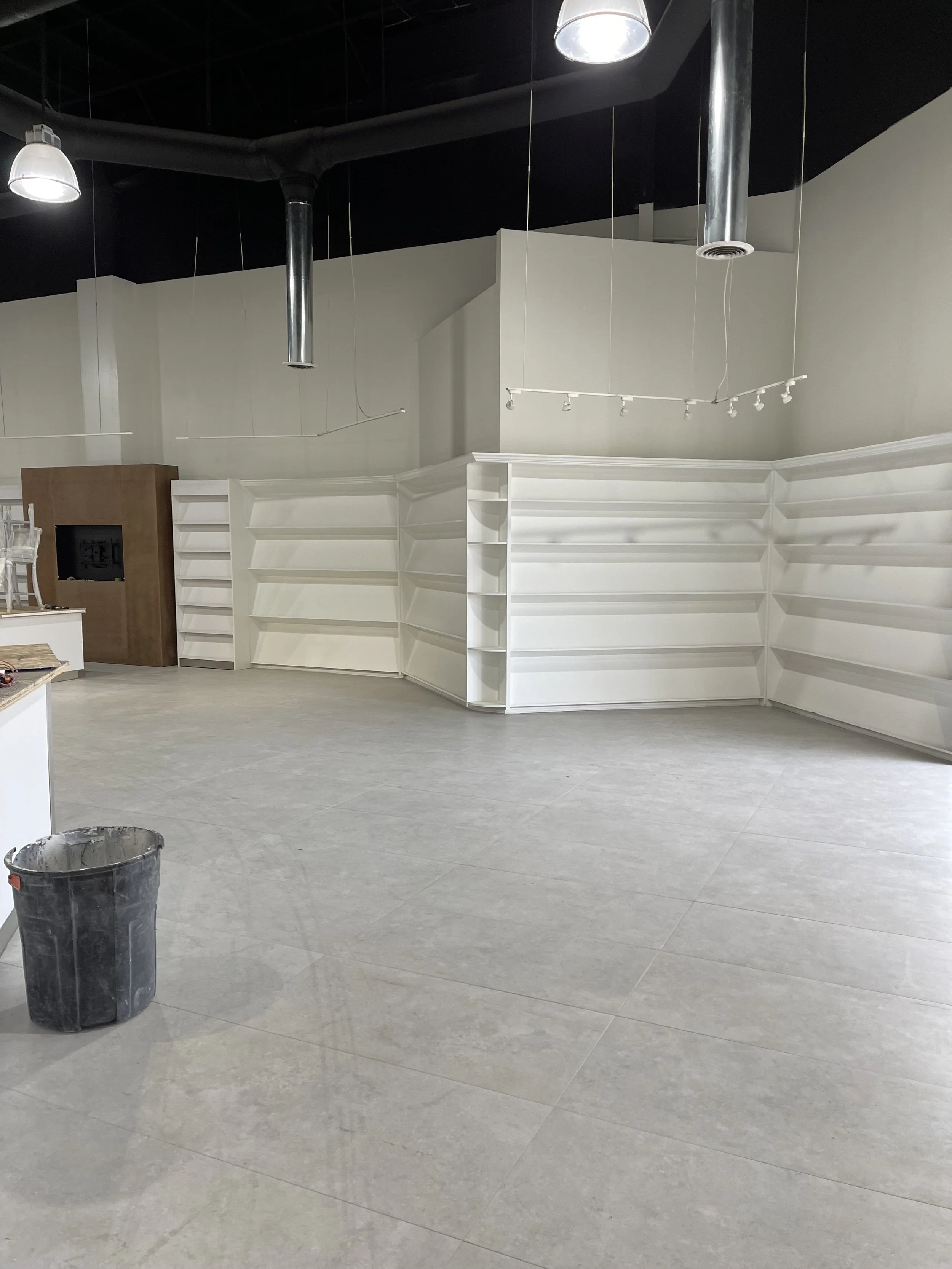 An interior of a store or showroom with white empty shelves, gray tile floors, and exposed black ceiling with pipes and lighting fixtures. A black trash can is in the foreground.