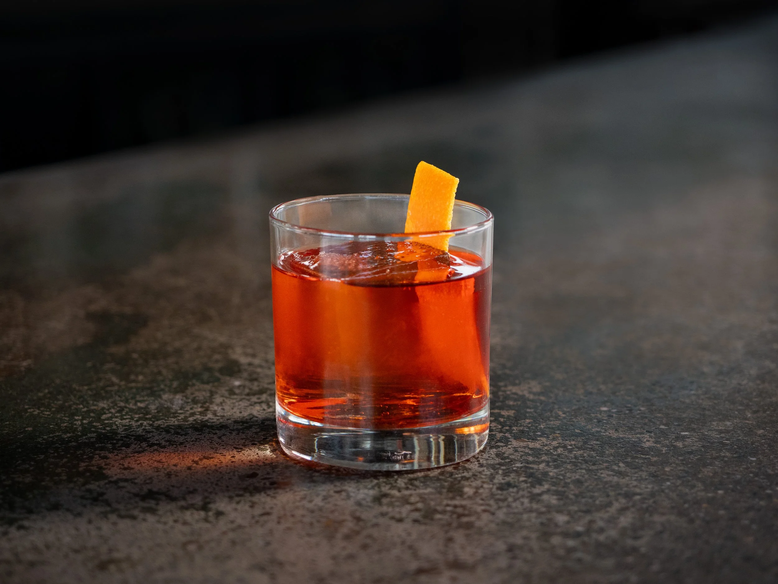 A clear glass of bright red beverage garnished with a rectangular orange strip on the rim, placed on a dark, textured surface with a black background.