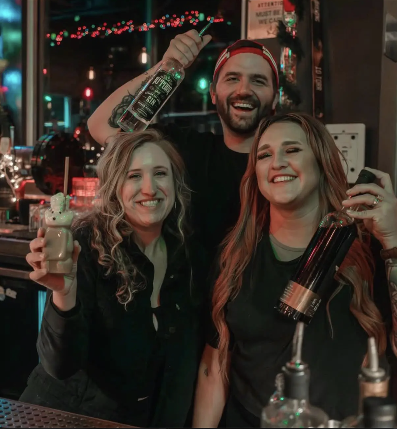 Three smiling people at a bar, each holding a drink. The man in the middle is wearing a red cap and holding a clear bottle, while the woman on the left holds a layered cocktail and the woman on the right holds a dark bottle. There are colorful string lights and bar equipment in the background.