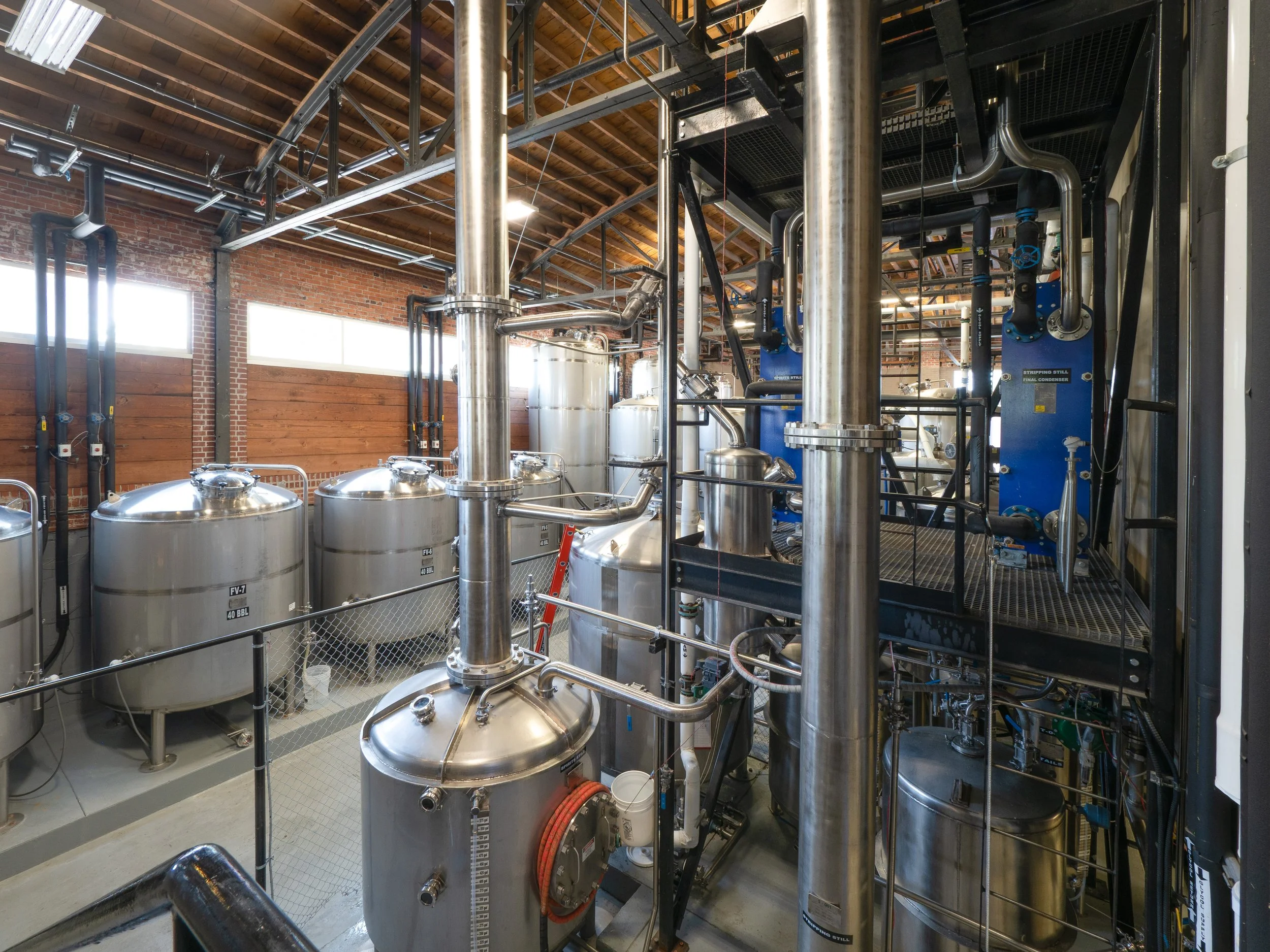 Industrial brewing equipment with stainless steel tanks, pipes, and valves inside a brick-walled building with wooden ceiling beams.